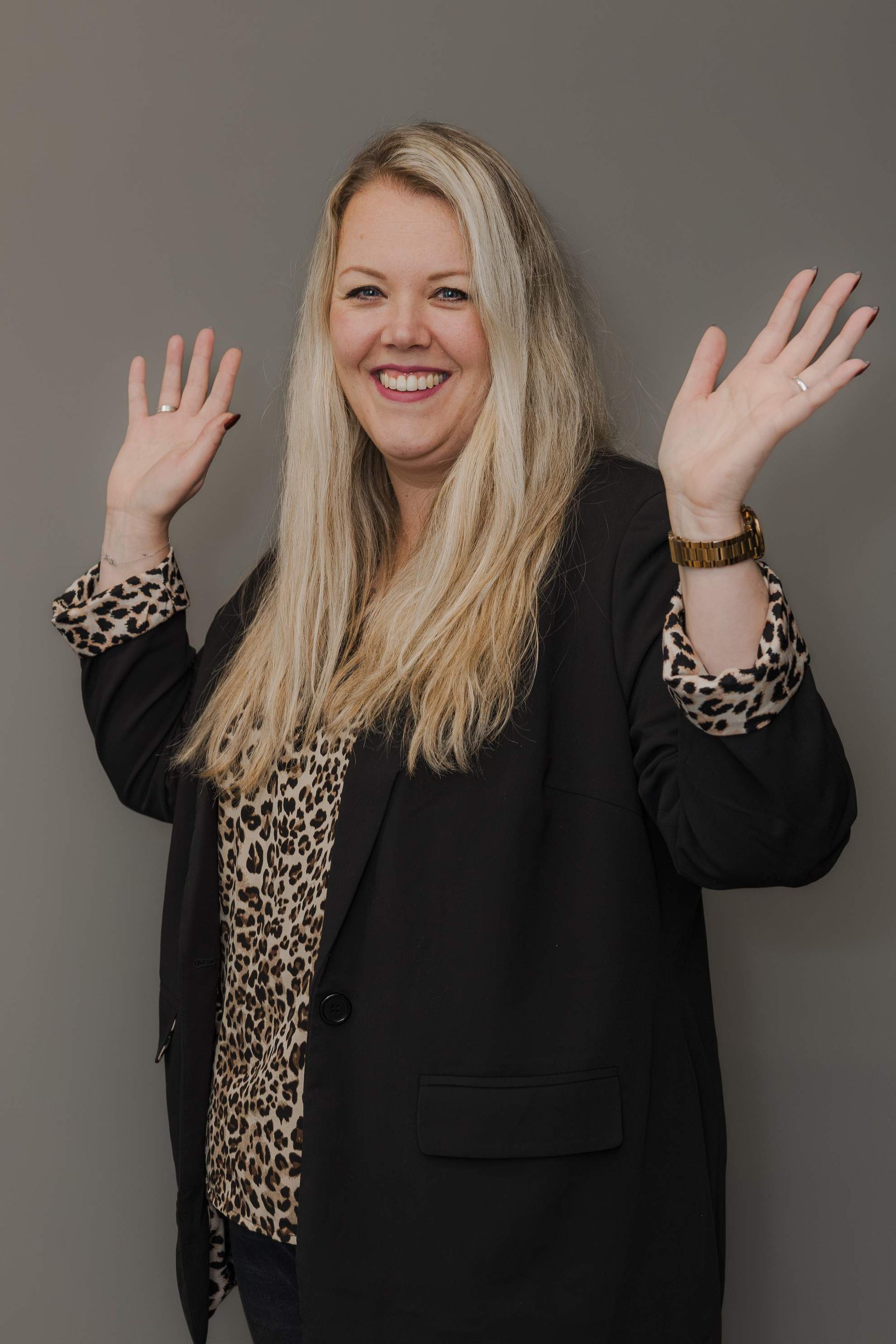 Vrouw met blond haar, armen omhoog, lachend, gekleed in een zwarte blazer met manchetten en blouse met luipaardprint.