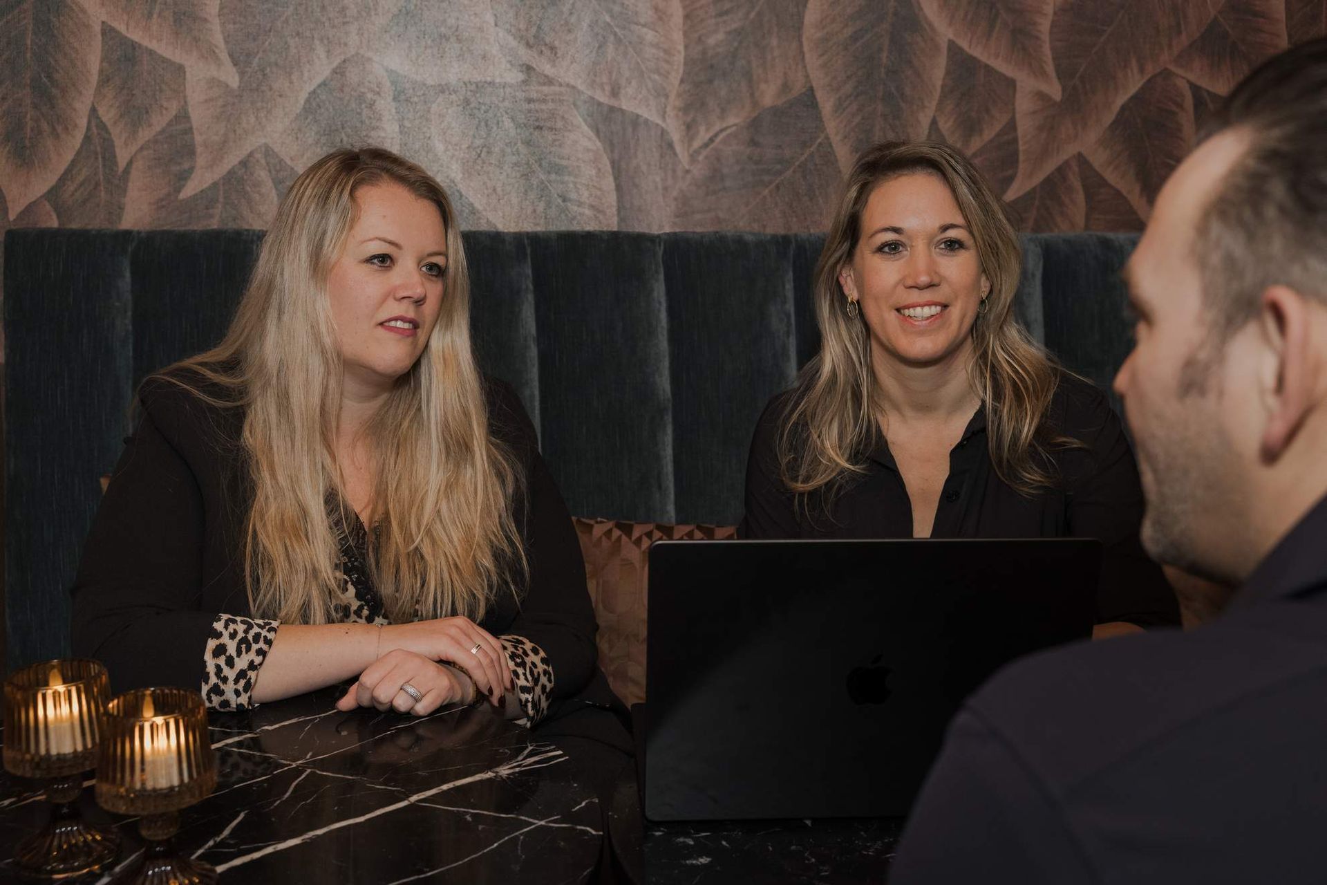 Twee vrouwen en een man zitten aan een tafel en kijken naar een laptop. De scène speelt zich af in een kamer.