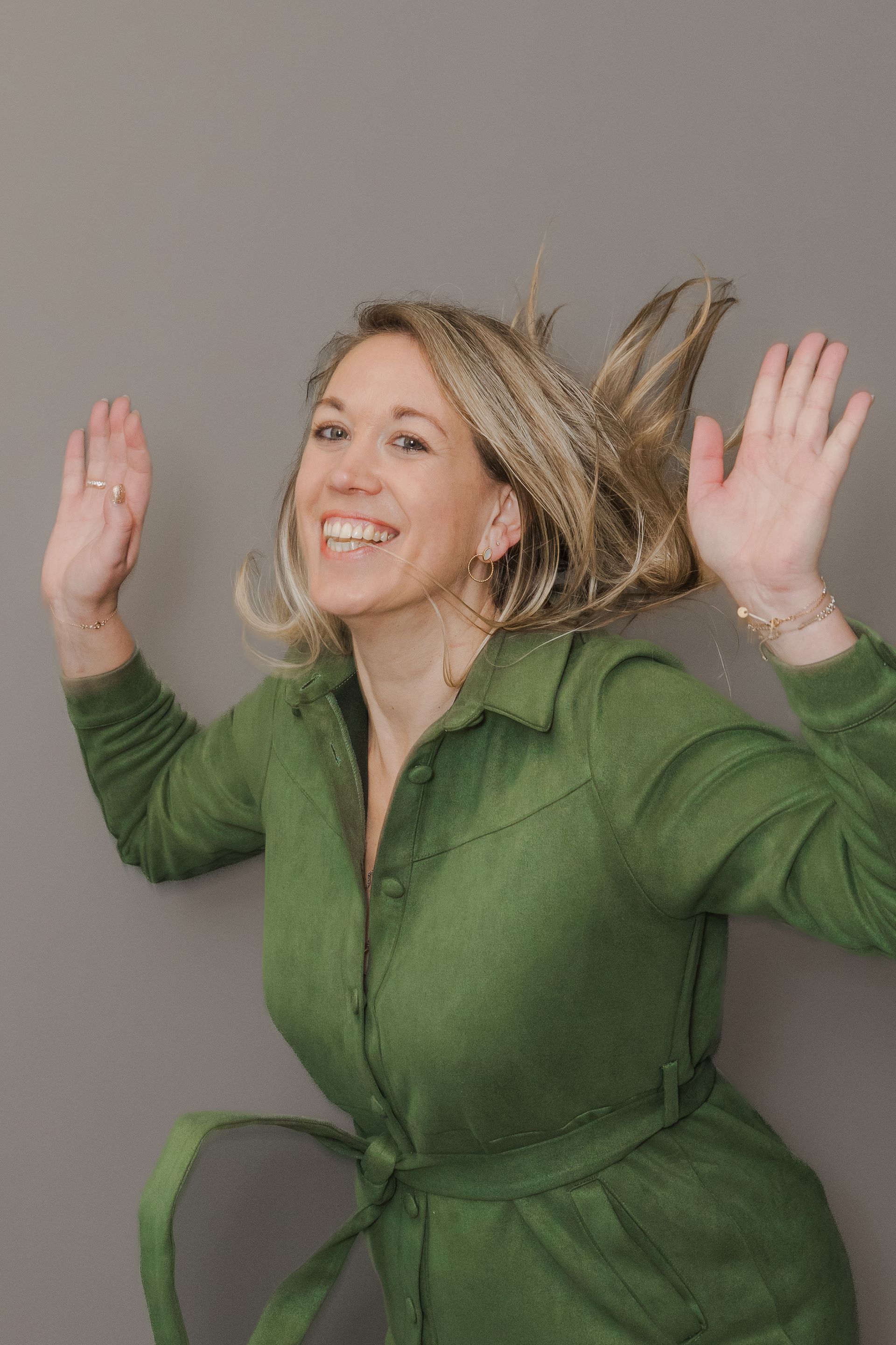 Vrouw in groene jumpsuit met opgeheven armen, lachend tegen een grijze muur.