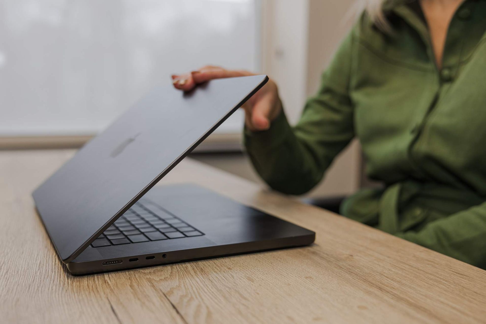 Iemand opent een donkere laptop op een licht houten bureau, binnenshuis.