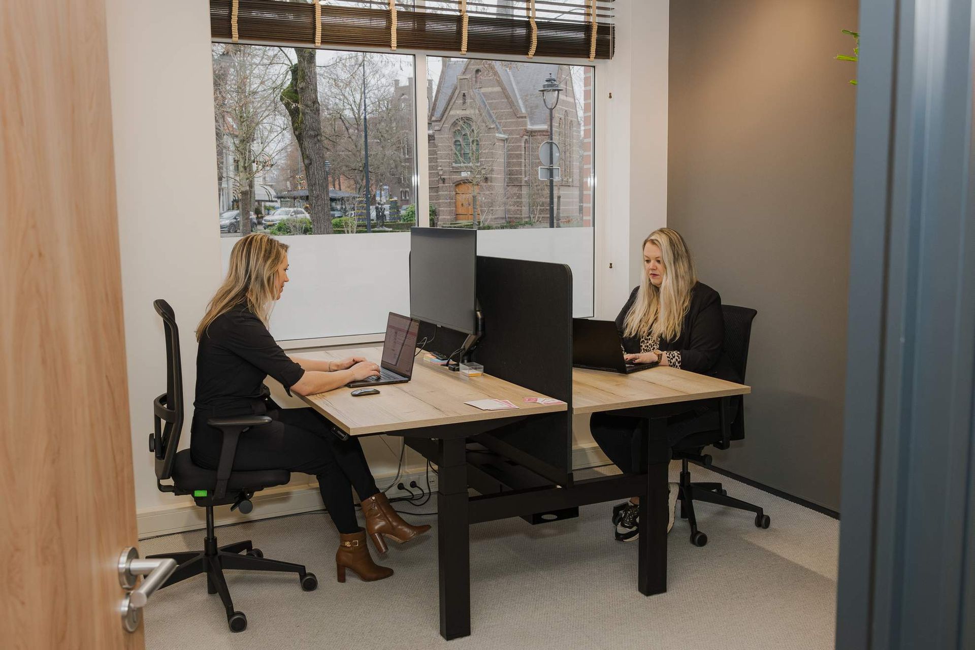 Twee vrouwen werken aan bureaus in een modern kantoor, met een raam en uitzicht op een kerk buiten.