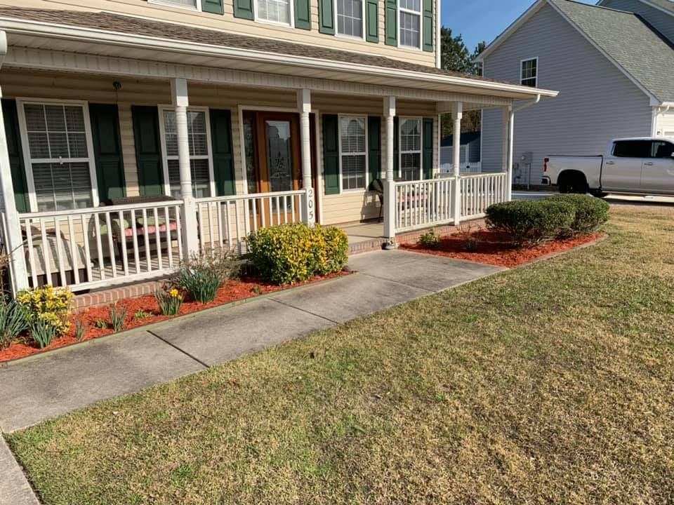 A house with a porch and in front of it is a landscaped garden bed with red mulch and small bushes.