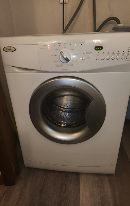 A white front-loading Whirlpool washing machine with a silver door, centered in a room with light wood-style flooring.
