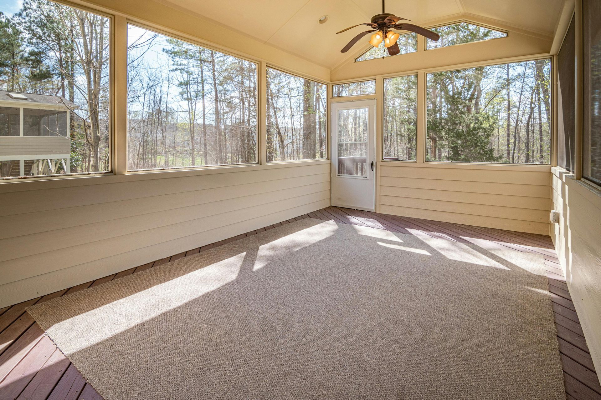 Screened-in porch with neutral-colored carpet, ceiling fan, and door; sunlight streams through windows into the woods.