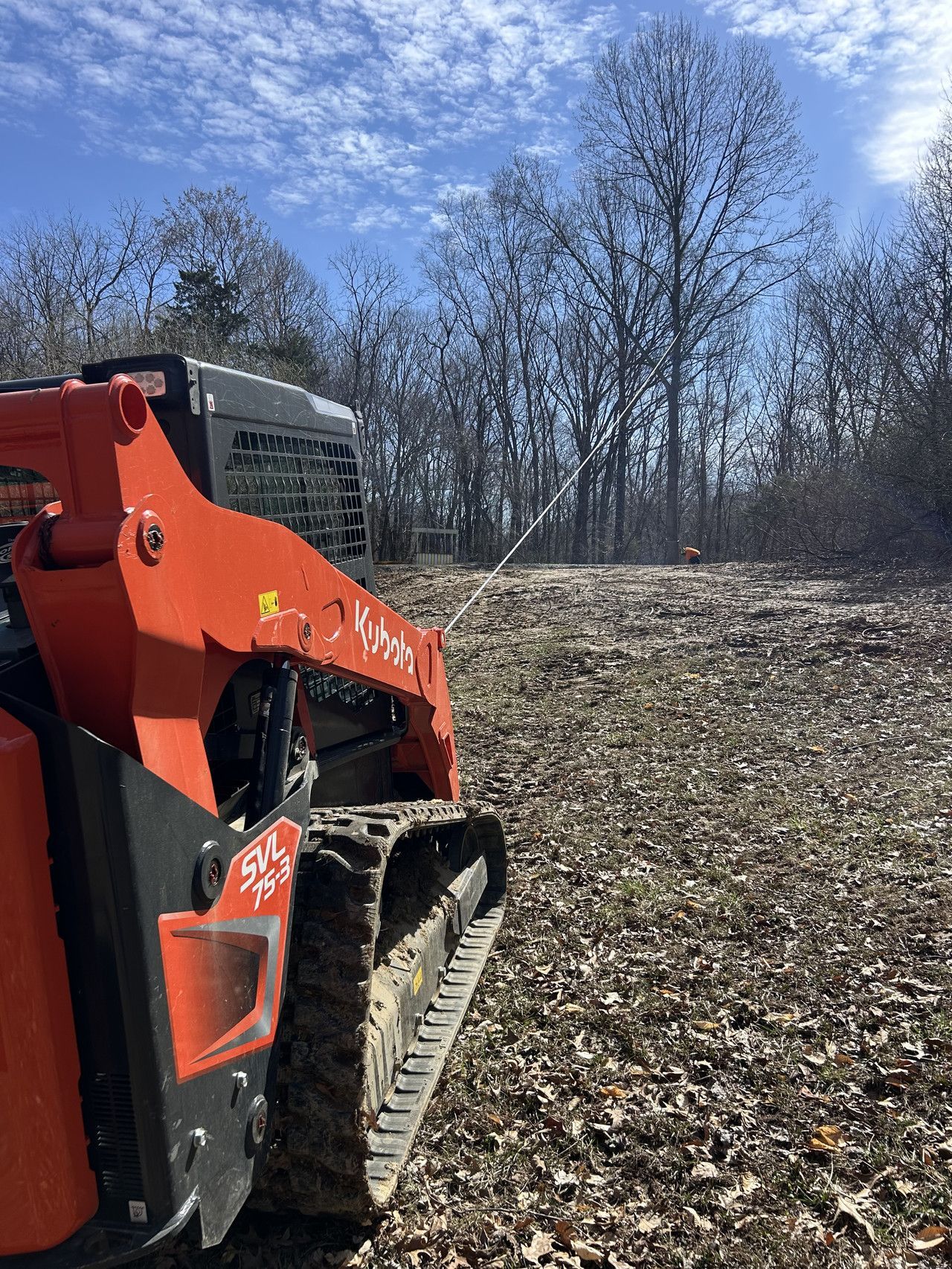 An orange Kubota track loader sits on a cleared, leaf-covered field with a line of trees in the background under a blue sky.