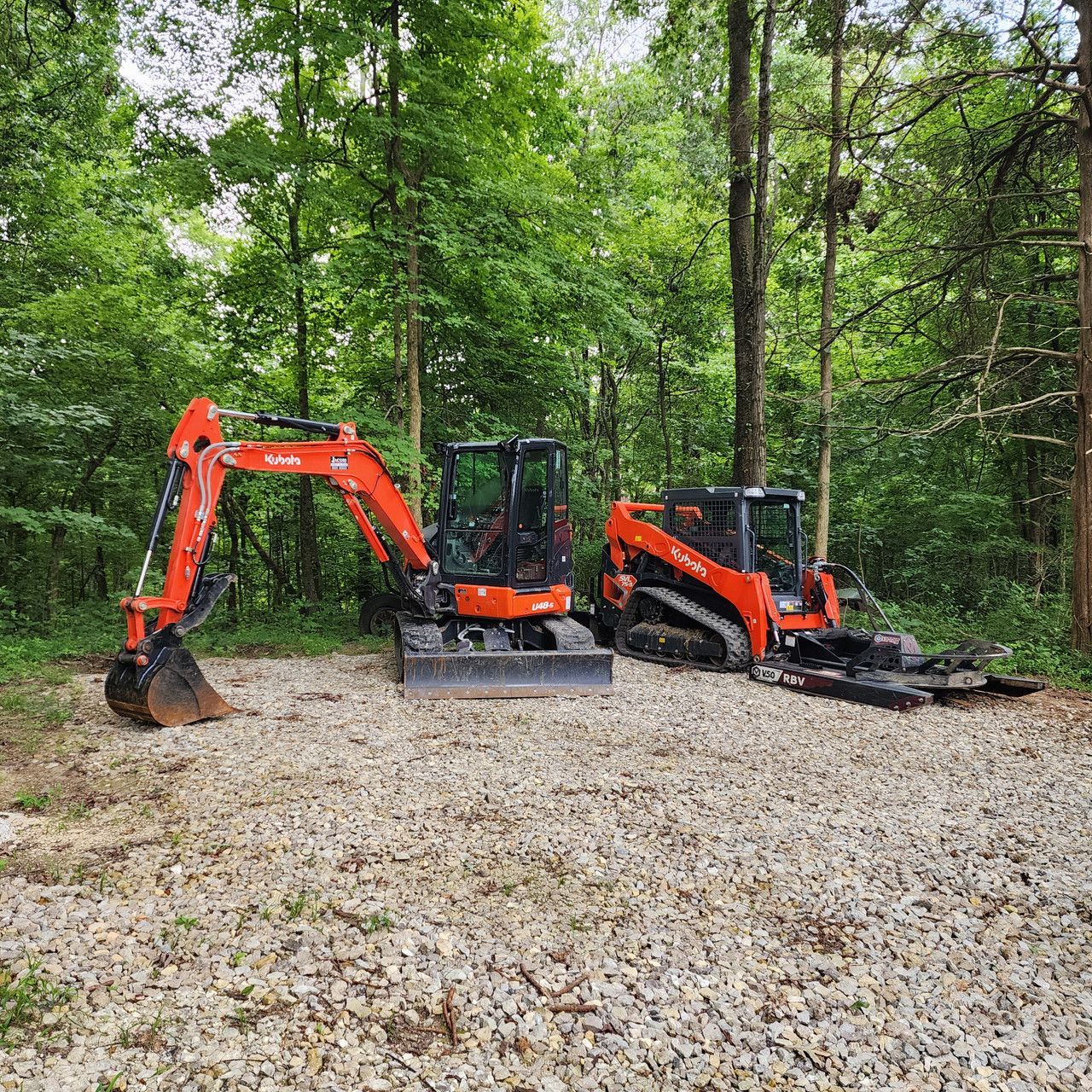 An orange excavator and an orange skid steer parked on a gravel clearing in a dense, green forest.