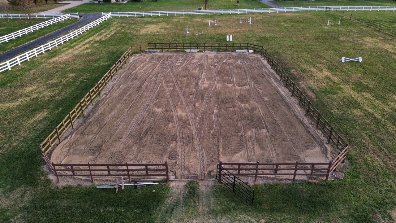 An aerial view of a rectangular, sandy horse riding arena enclosed by a wooden fence in a grassy field.