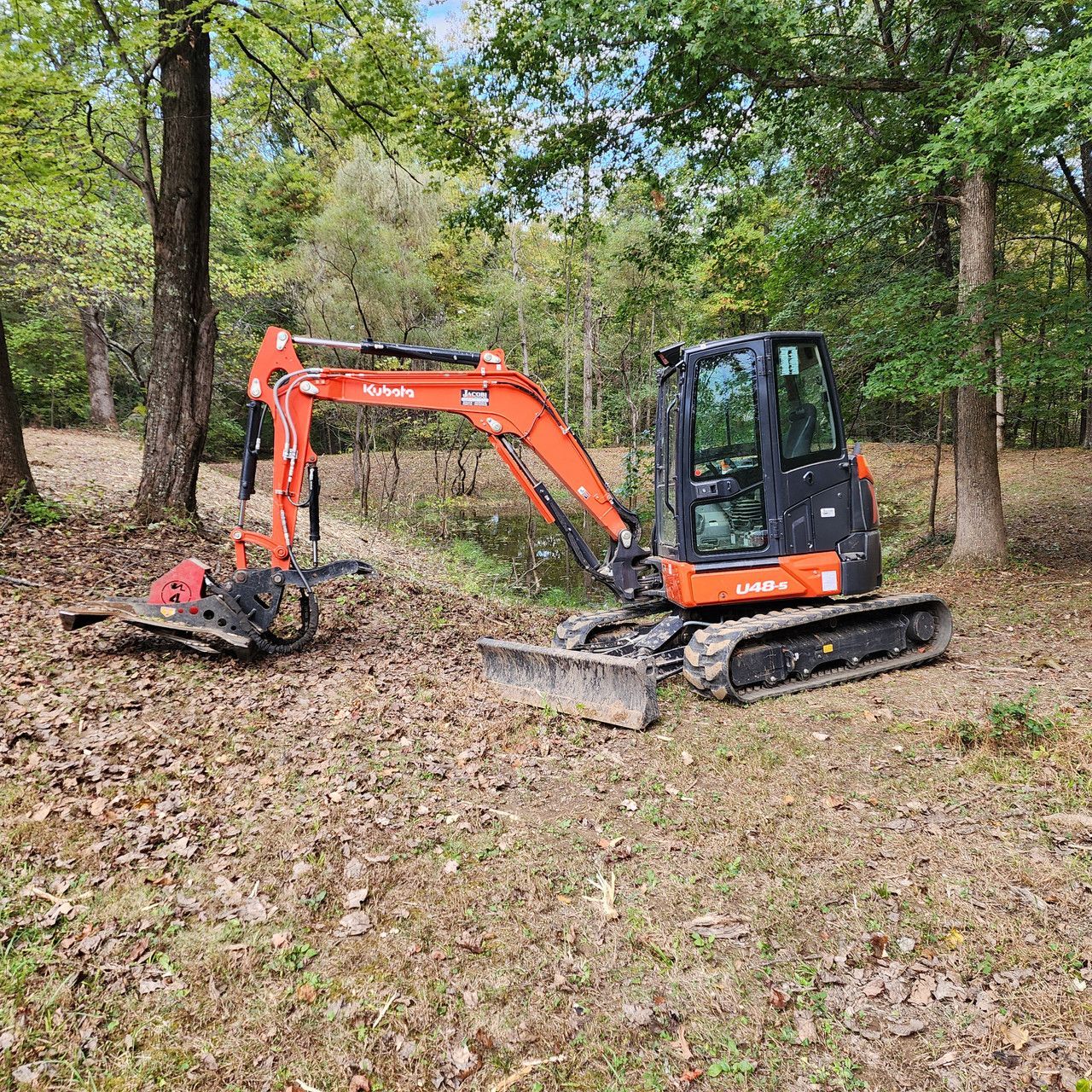 A bright orange Kubota mini excavator with a brush cutter attachment sits in a wooded area covered in fallen leaves.