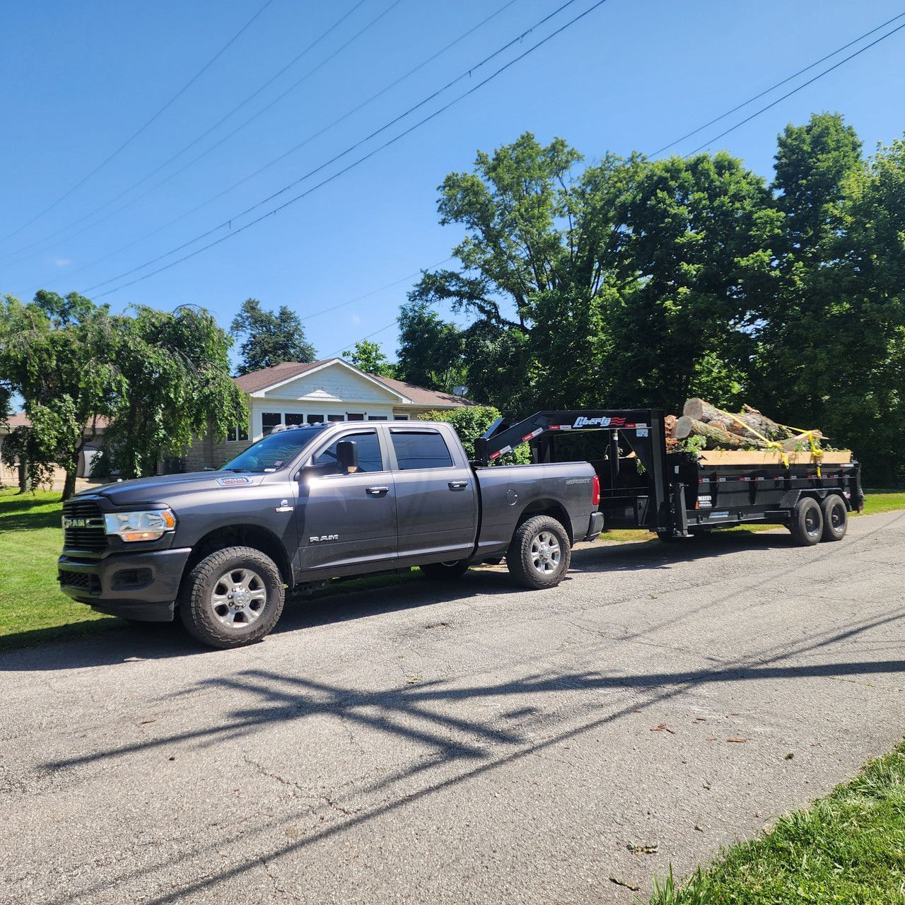A grey Ram pickup truck parked on an asphalt driveway, pulling a flatbed trailer loaded with cut tree logs.