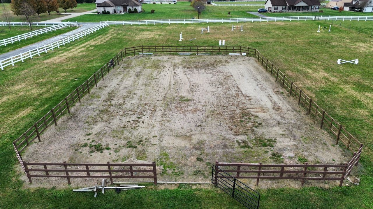 An aerial view of a fenced, dirt horse riding arena in a grassy field, with residential houses in the background.