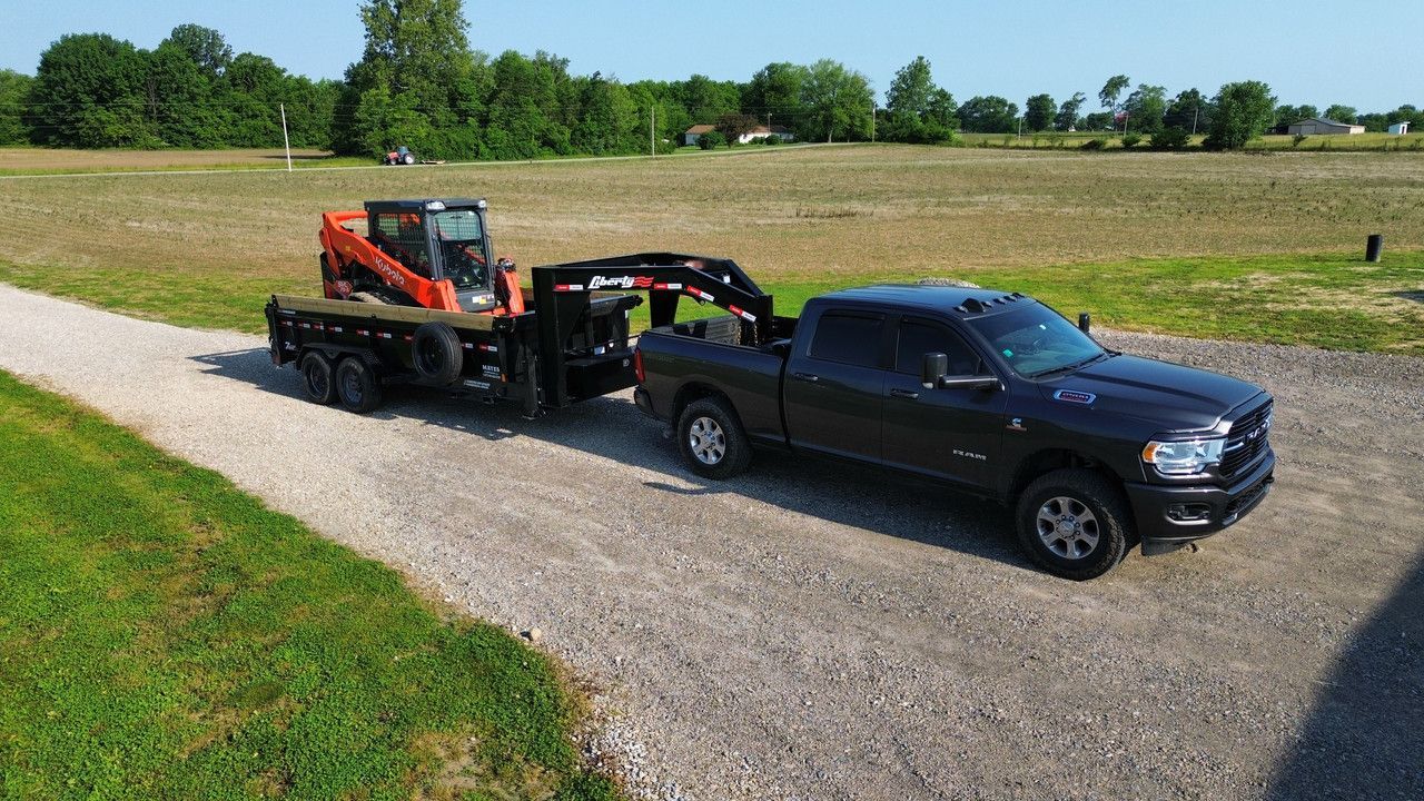 A dark pickup truck tows a black flatbed trailer carrying an orange skid-steer loader on a gravel driveway in a field.