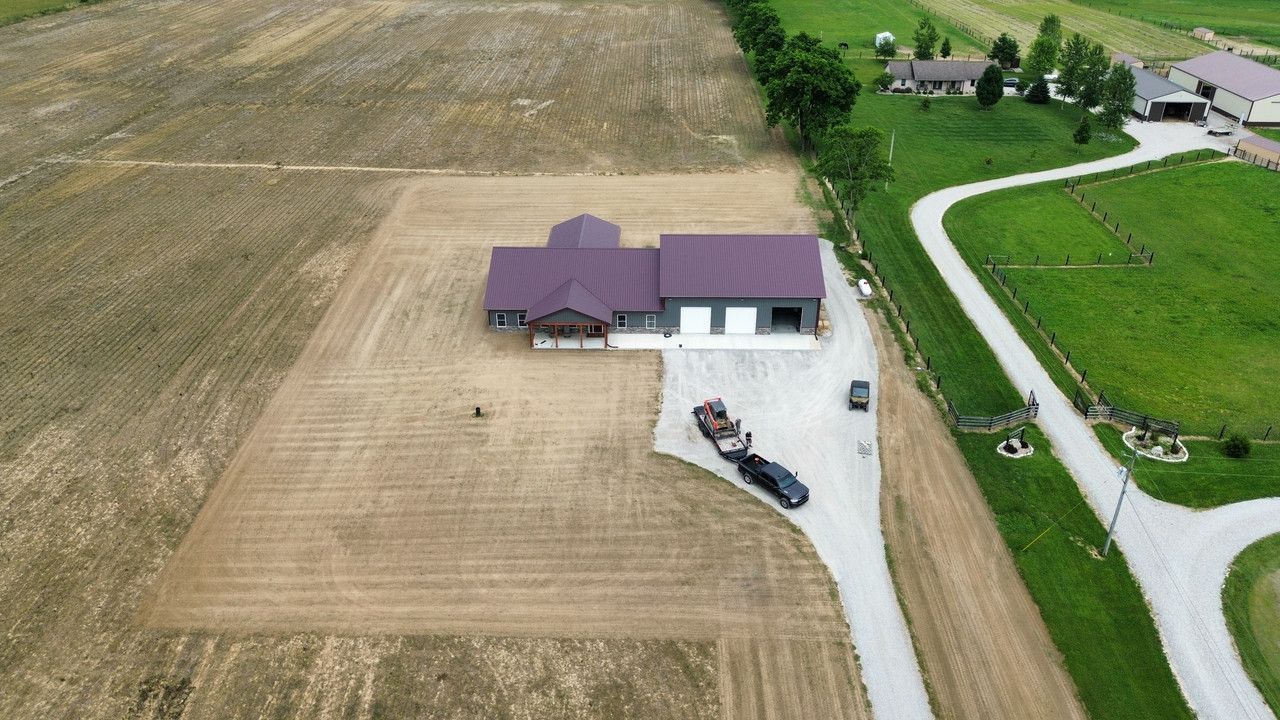 An aerial view of a farmhouse with a purple roof, surrounded by grassy fields and a long gravel driveway.