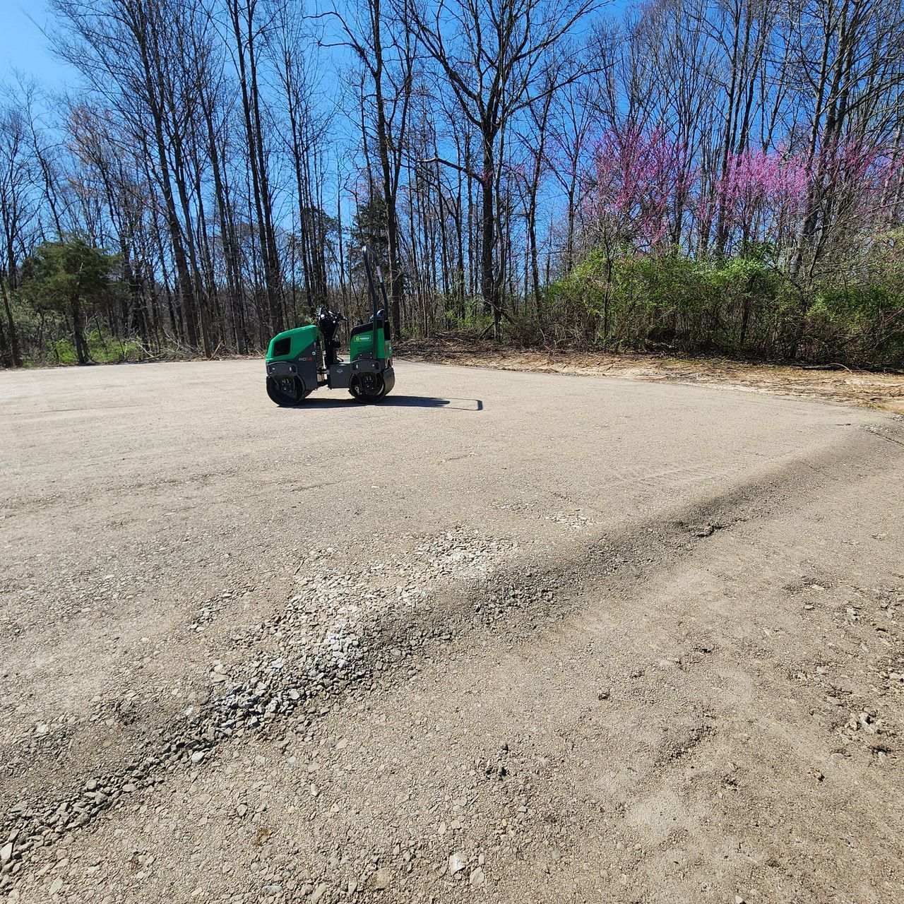 A green industrial roller sits on a level gravel lot in front of a wooded area with budding spring trees.