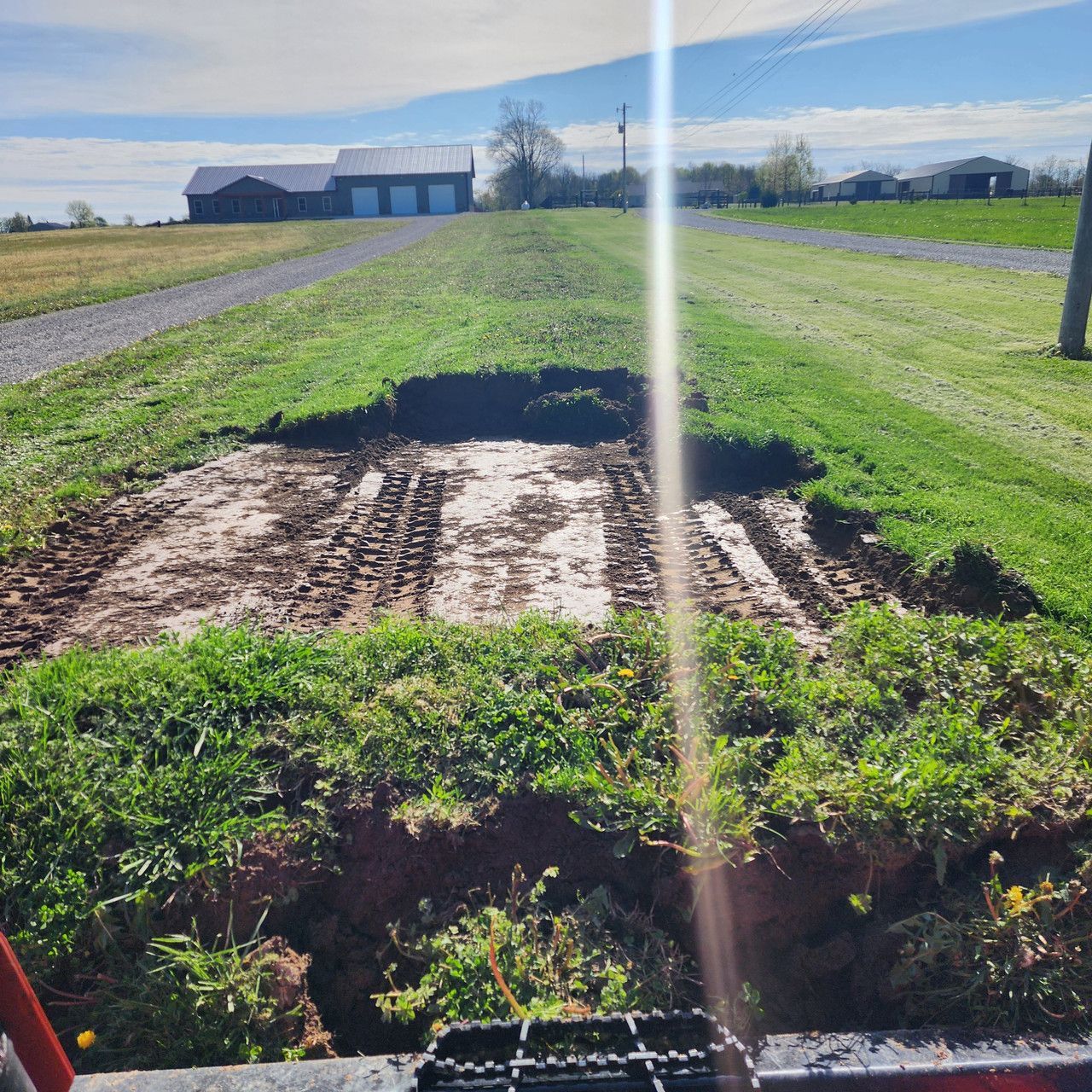 A large, rectangular section of turf has been removed from a lawn, revealing mud and tire tracks, near a rural farm area.