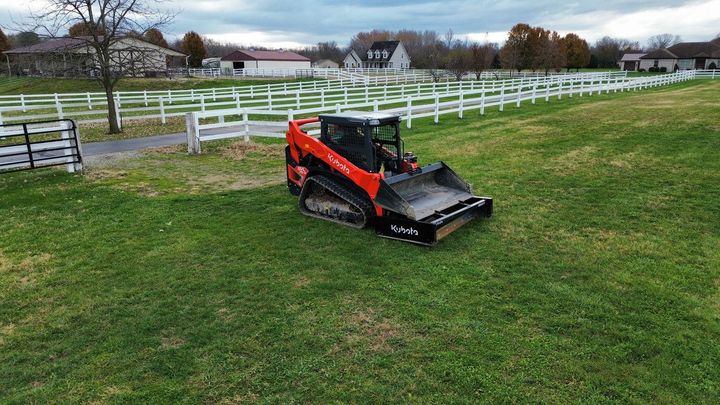 An orange Bobcat compact track loader with a brush cutter attachment sits on a grassy field by a white fence.