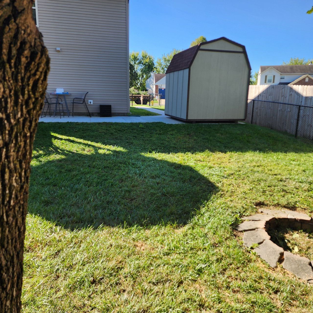 A backyard scene featuring a tan house, a small storage shed, a lawn, and a stone-lined fire pit area in the foreground.