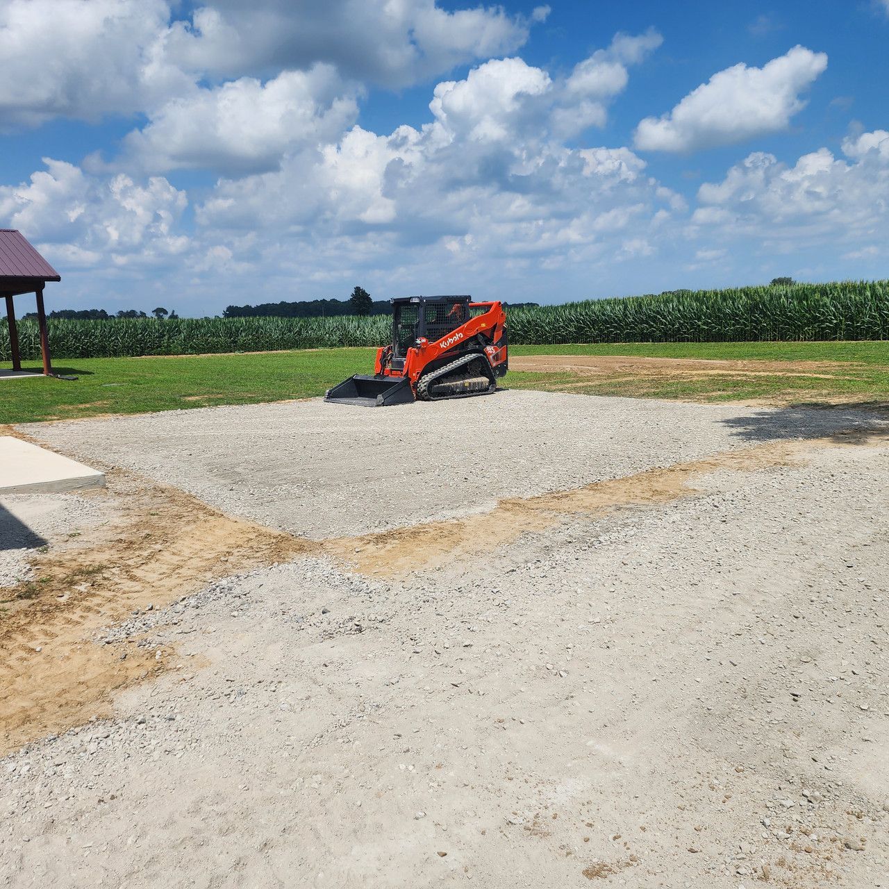 An orange skid steer loader sits on a leveled gravel pad in a field under a cloudy blue sky.