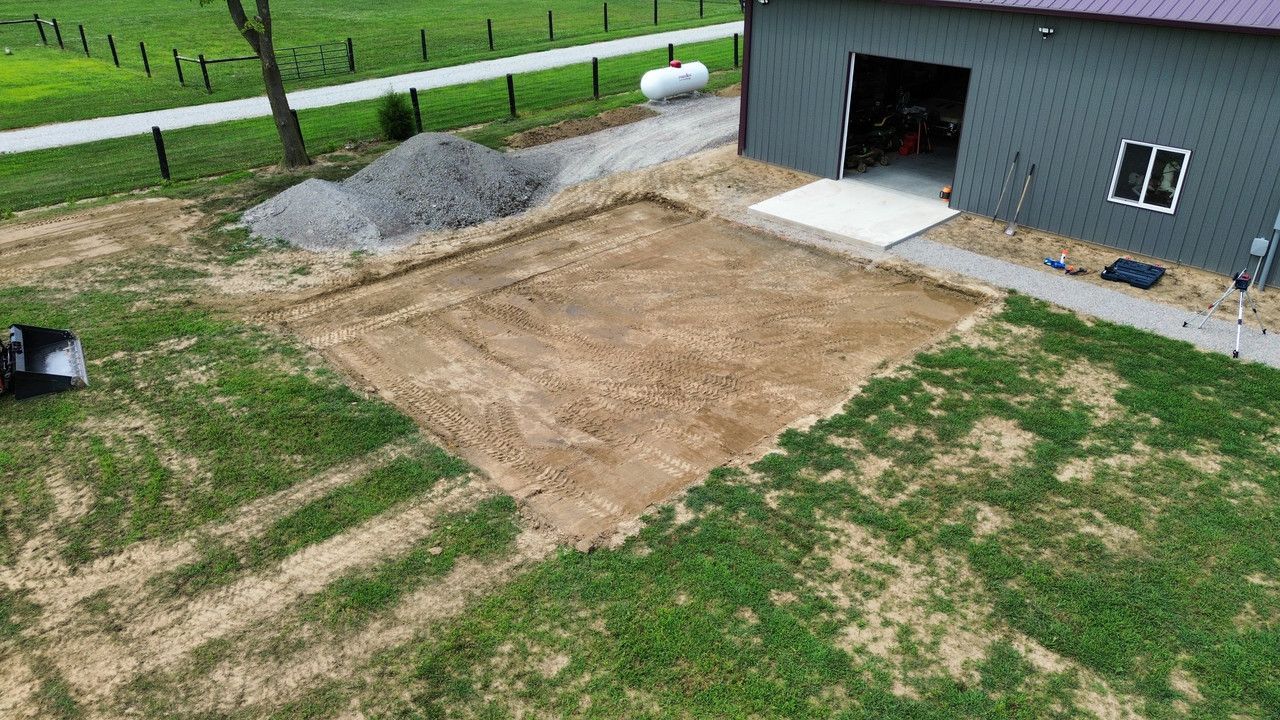 An aerial view of a site prepared for construction next to a grey metal building, featuring a cleared, level soil area.