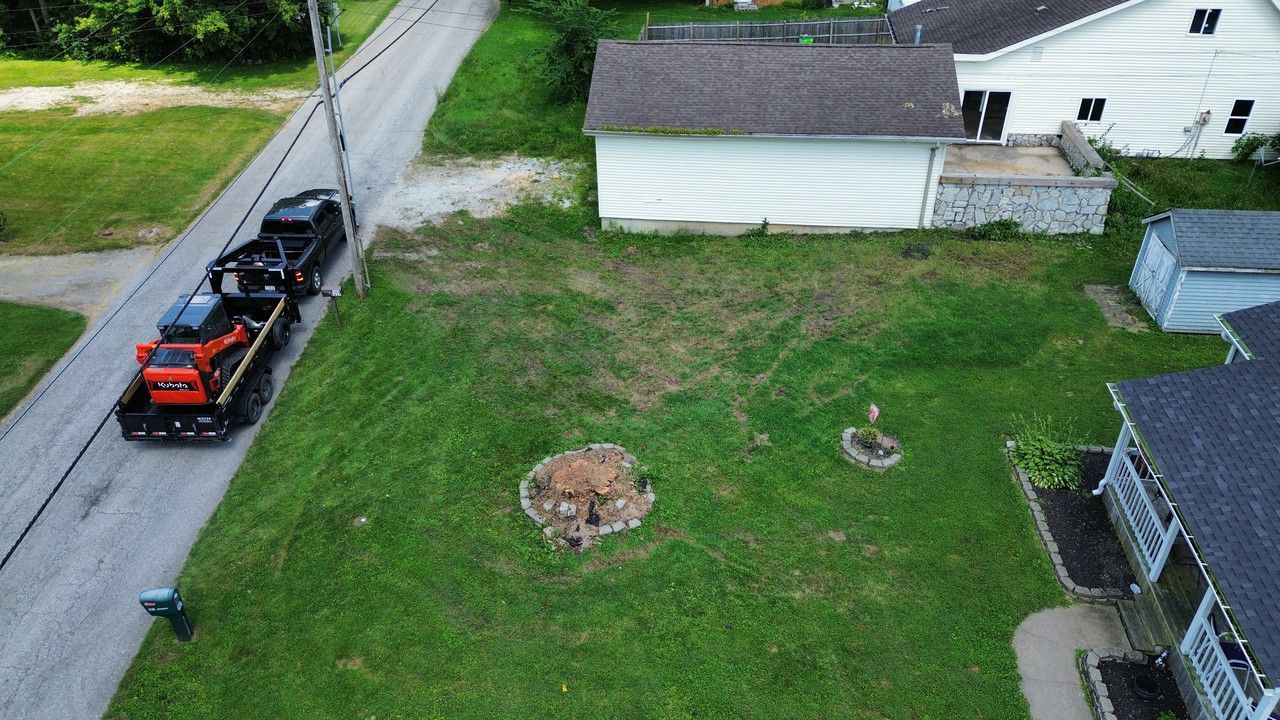 An aerial view of a grass yard with two tree stumps, next to a street where a truck is towing a piece of orange equipment.