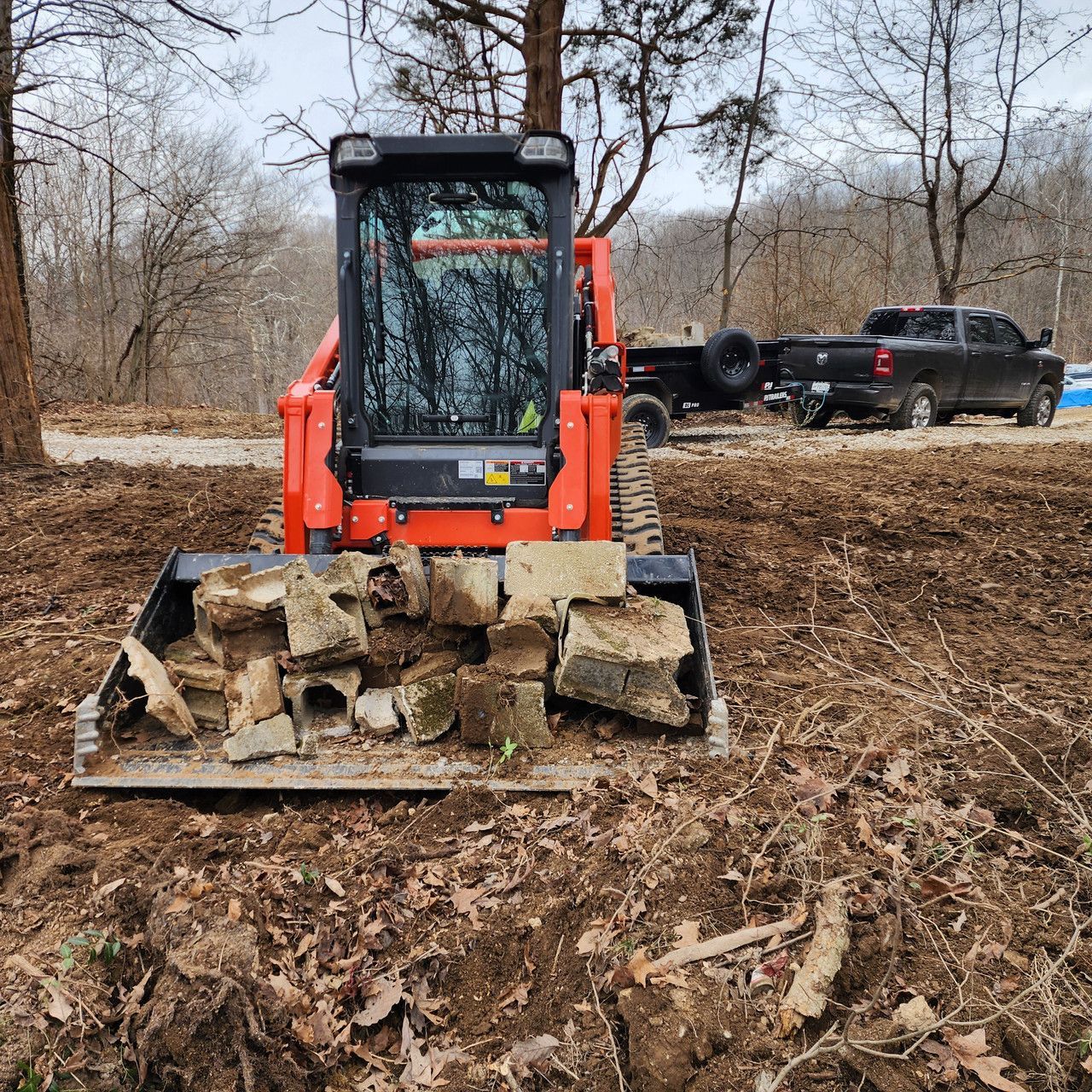 An orange skid steer carrying a bucket full of concrete rubble in a wooded, muddy field with a truck parked nearby.