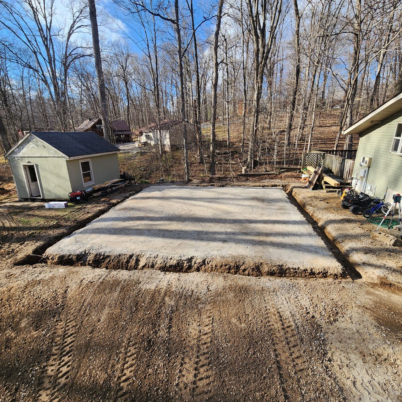 A level, rectangular gravel base for a building foundation sits in a wooded yard between two small structures.