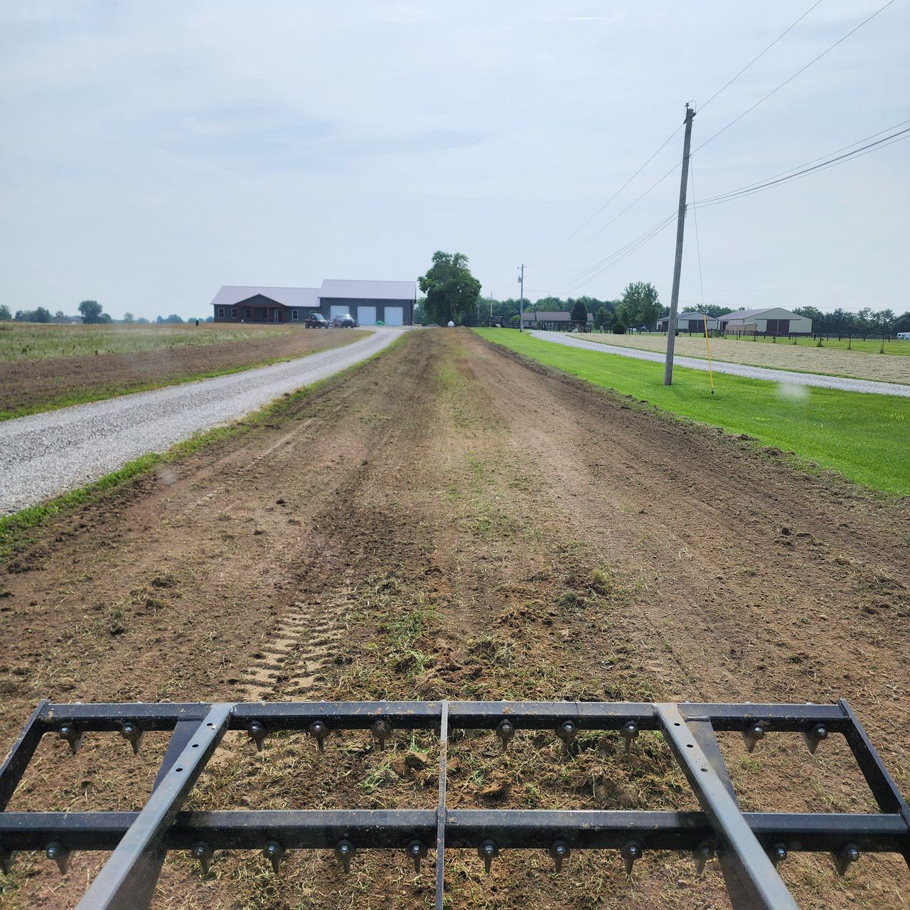 A first-person view from a tractor looking out over a tilled field toward a rural property with a house and garage.