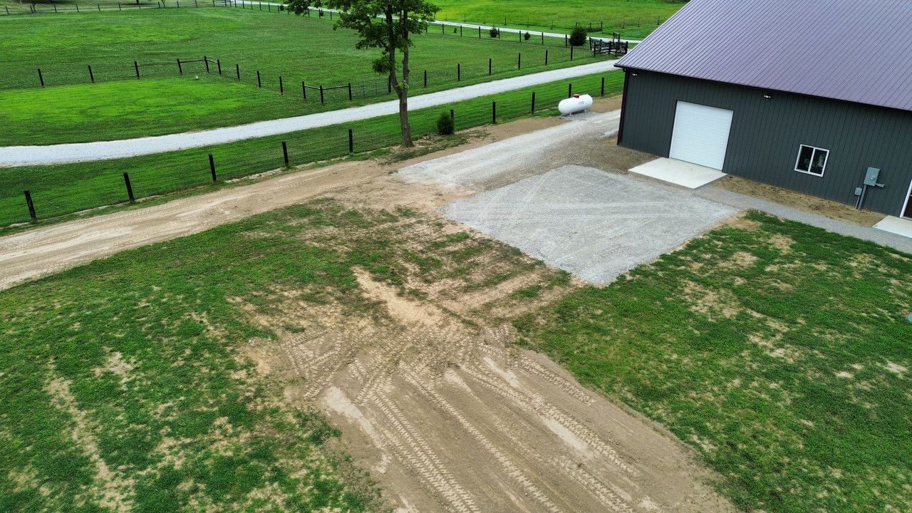 An aerial view of a gray barn with a gravel area in front, adjacent to a dirt path and a green grass field.