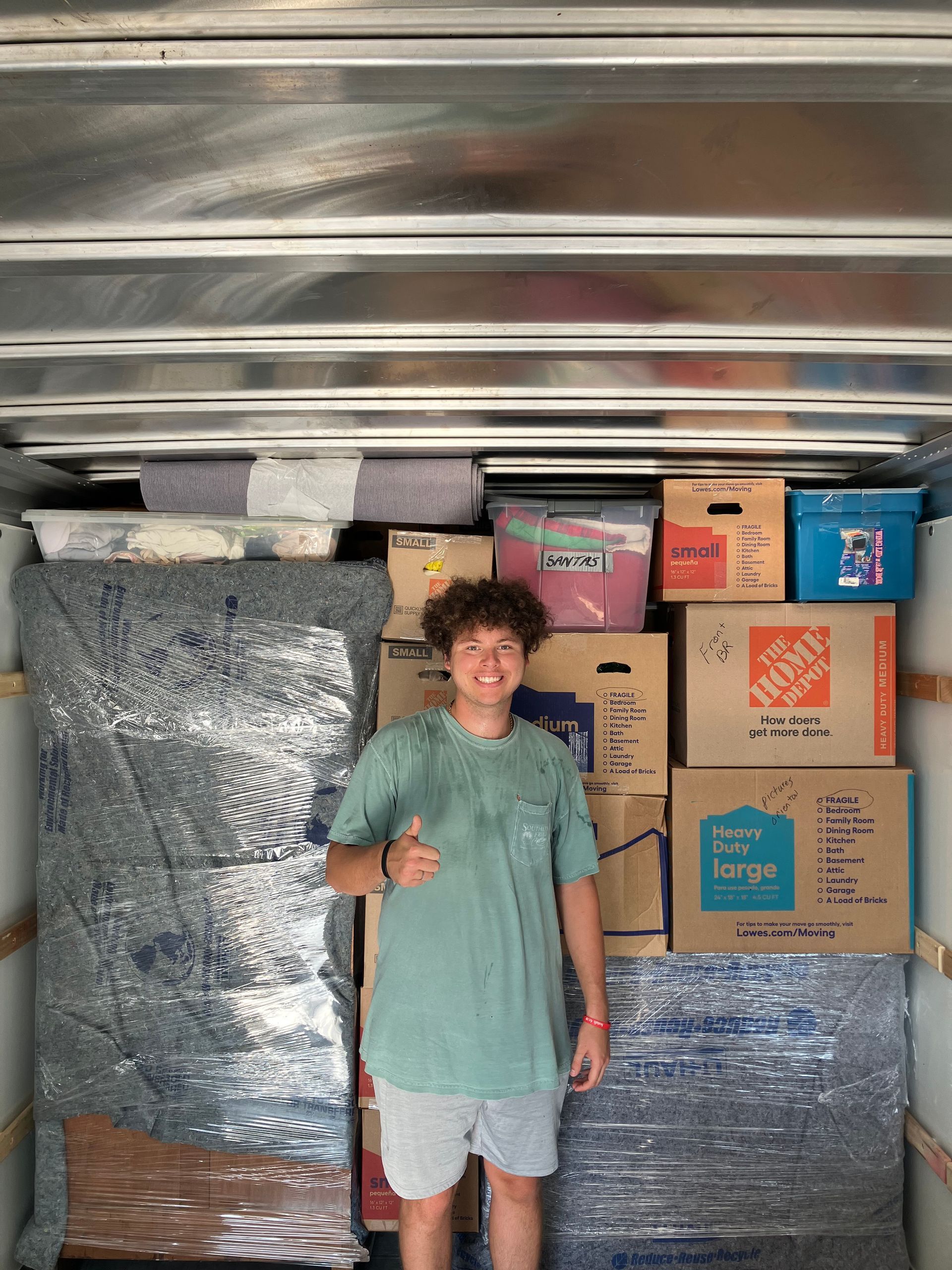 A young man is standing in a truck filled with boxes and giving a thumbs up.