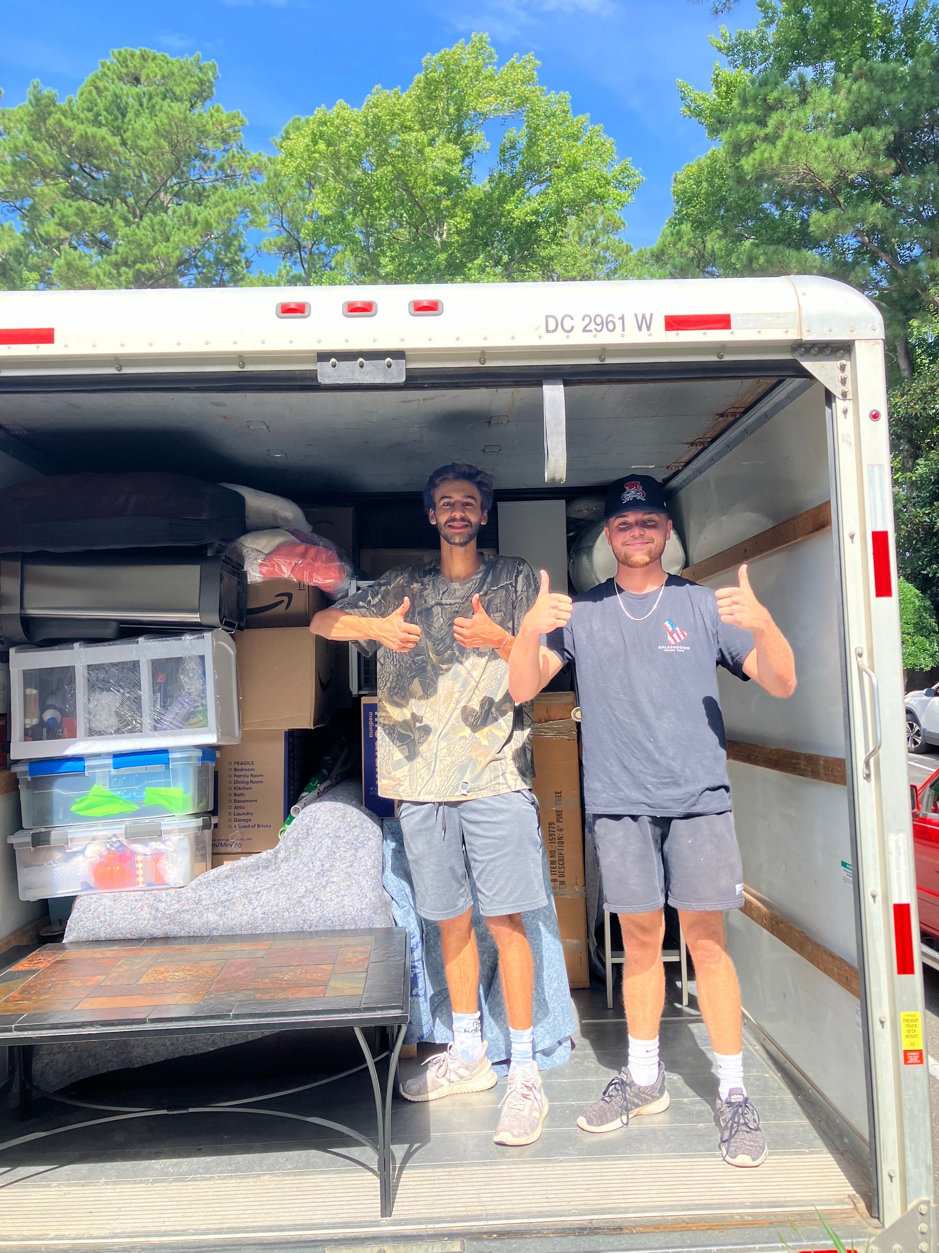 Two men are standing in the back of a moving truck giving a thumbs up.