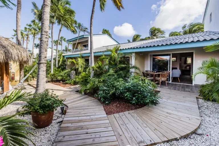 Wooden walkway through tropical foliage, leading to a patio with a table and chairs, near a cream-colored building.