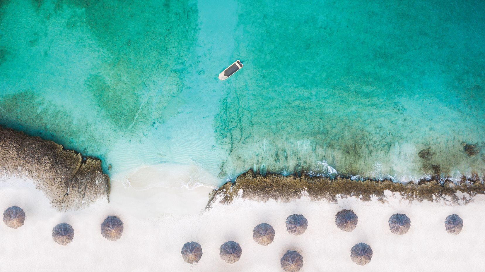 Aerial view of a white sandy beach with turquoise water. A small boat floats offshore, and beach umbrellas line the shore.