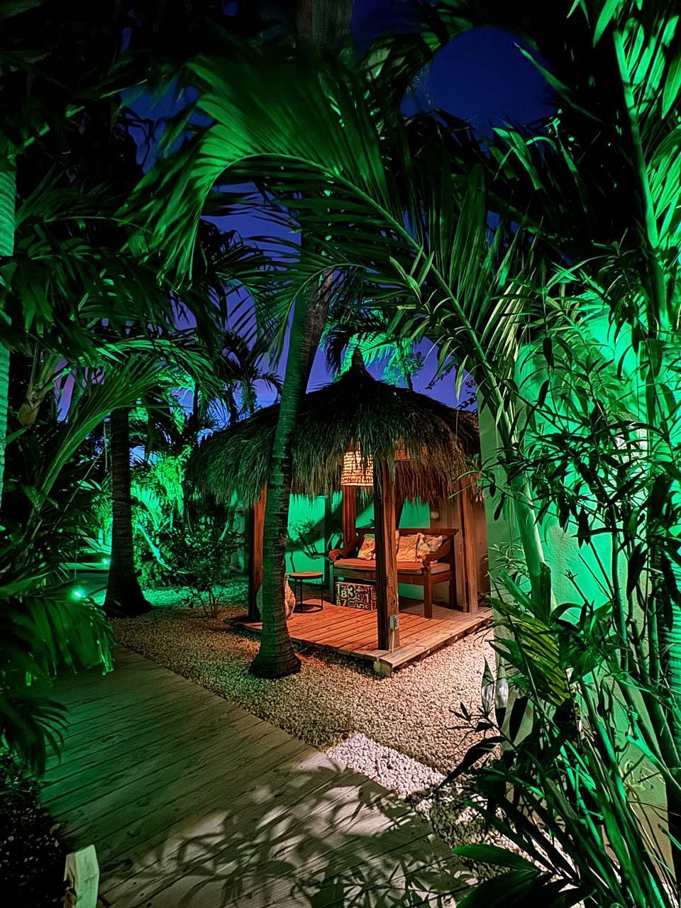 Nighttime tropical scene with a thatched-roof gazebo lit from within, surrounded by palm trees and green lighting.