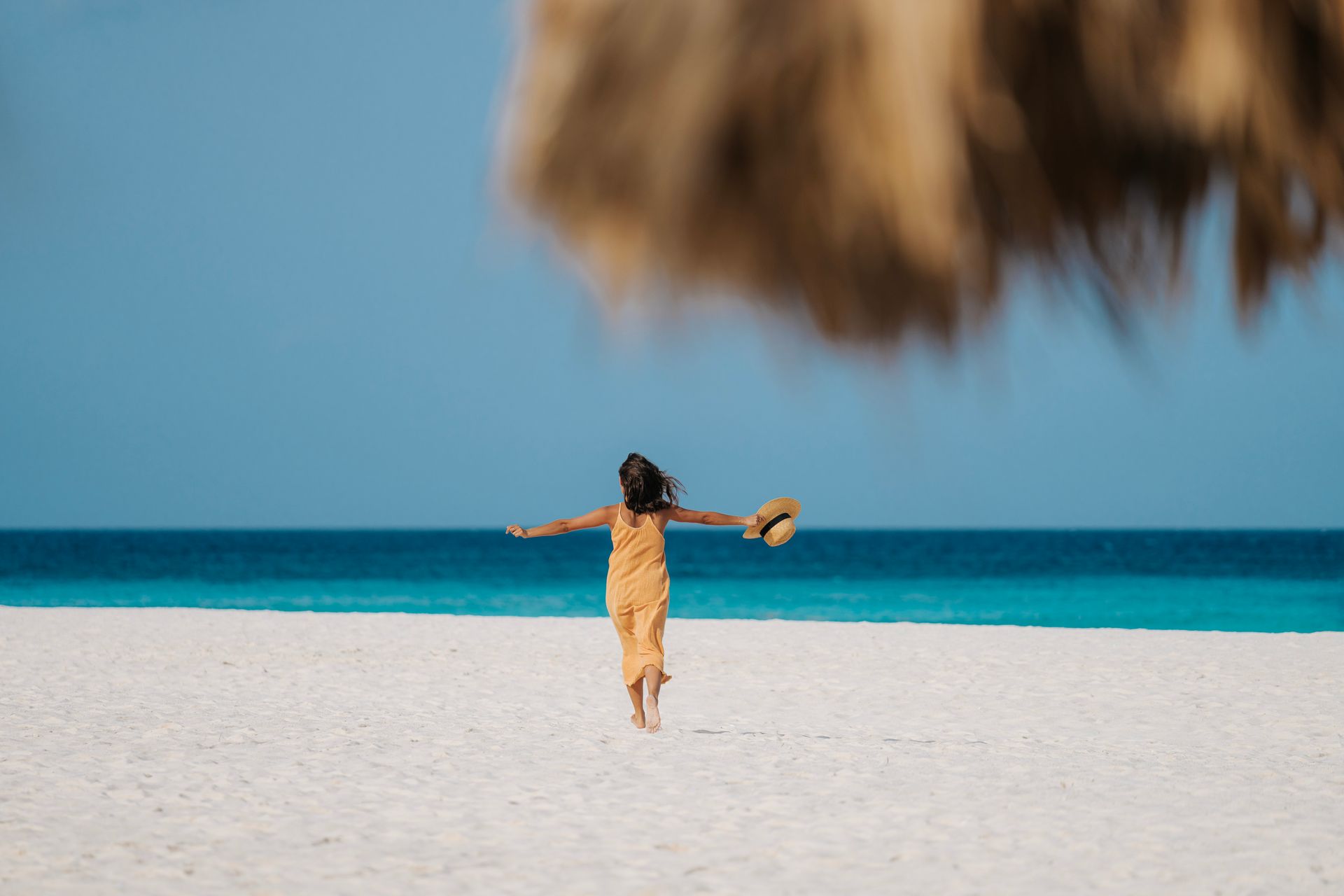 Woman runs on white sand toward turquoise water, holding a hat. Blue sky.