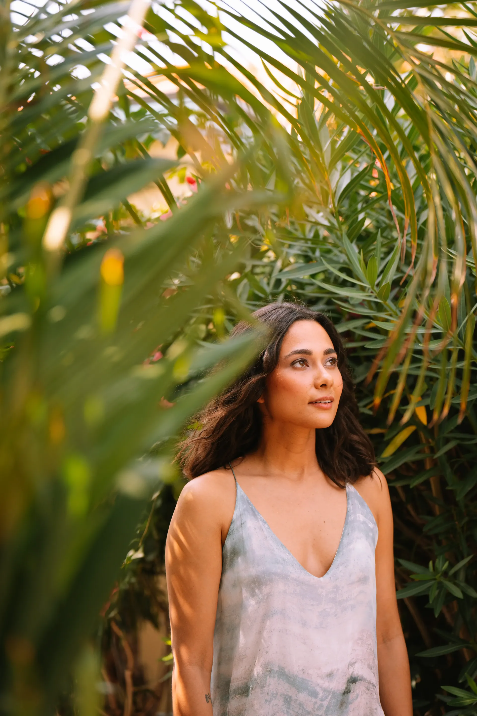 Woman in a light blue dress stands among green foliage, looking upward with a thoughtful expression.