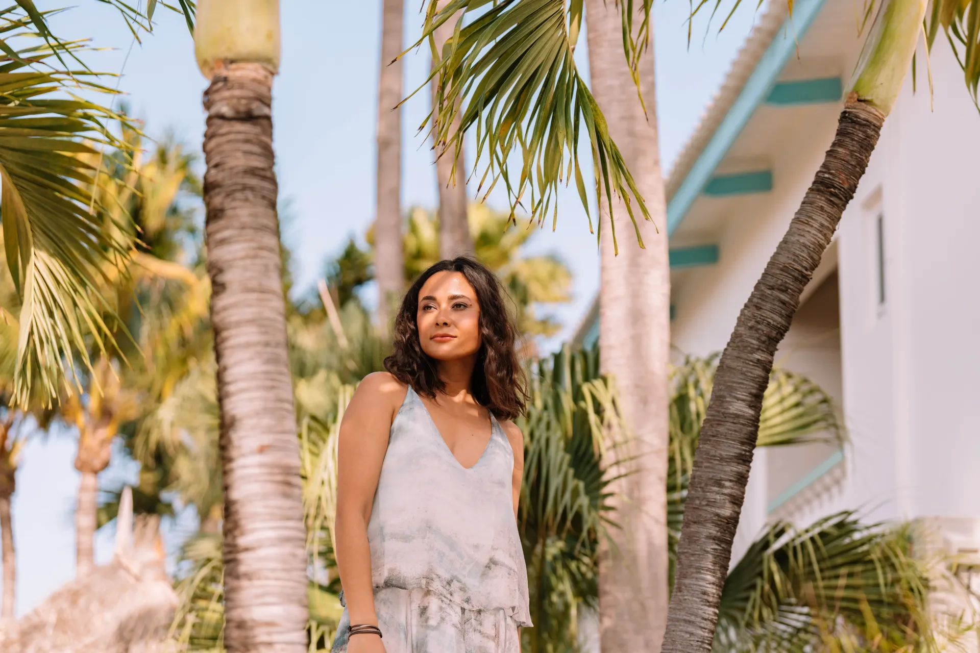 Woman in a white dress stands among palm trees in front of a white building with blue trim.