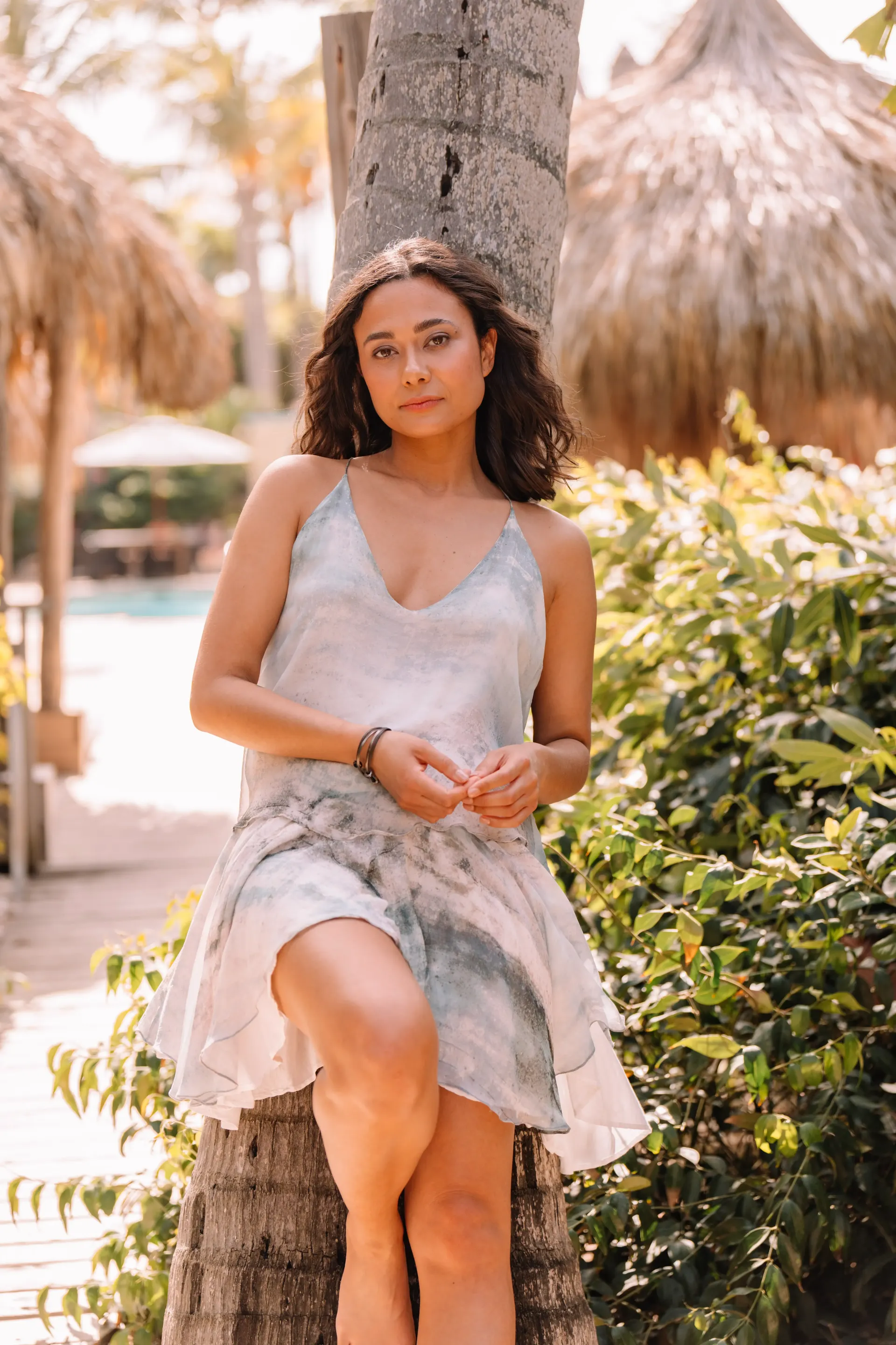 Woman in a flowing dress leans against a palm tree outdoors, near a thatch roof structure.