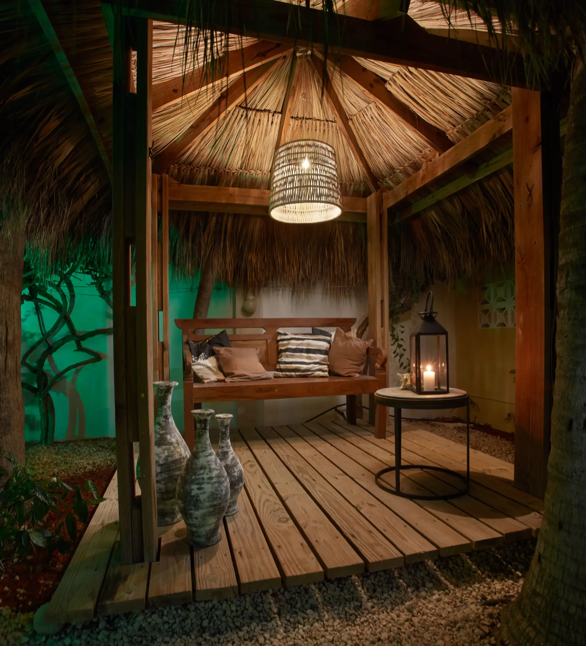 Wooden gazebo with thatched roof, bench, pillows, side table with lantern, and decorative vases.