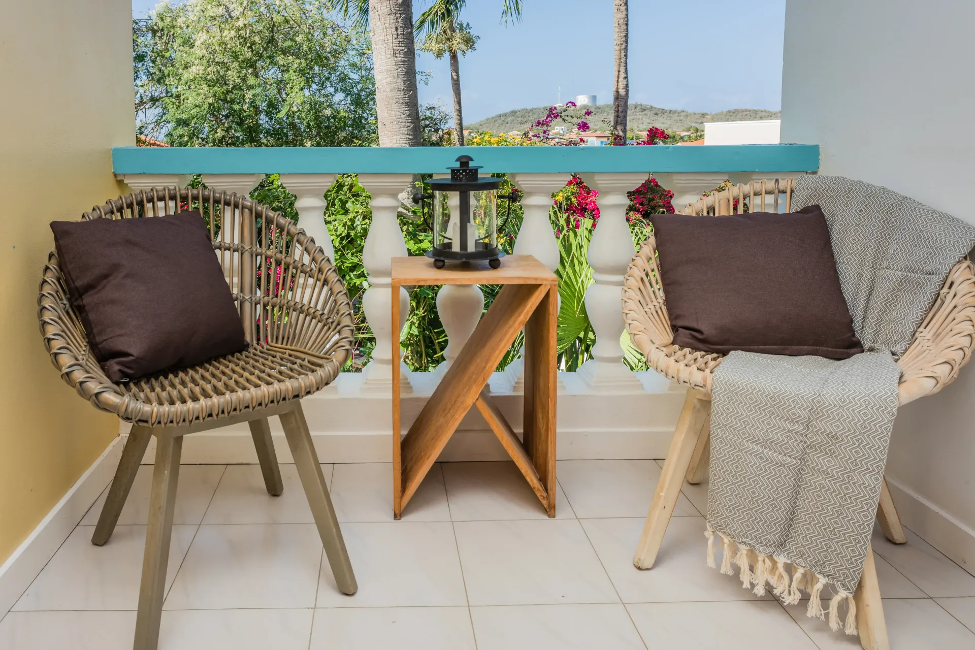 Two wicker chairs on a balcony with cushions and a blanket, a wooden side table, and a lantern.