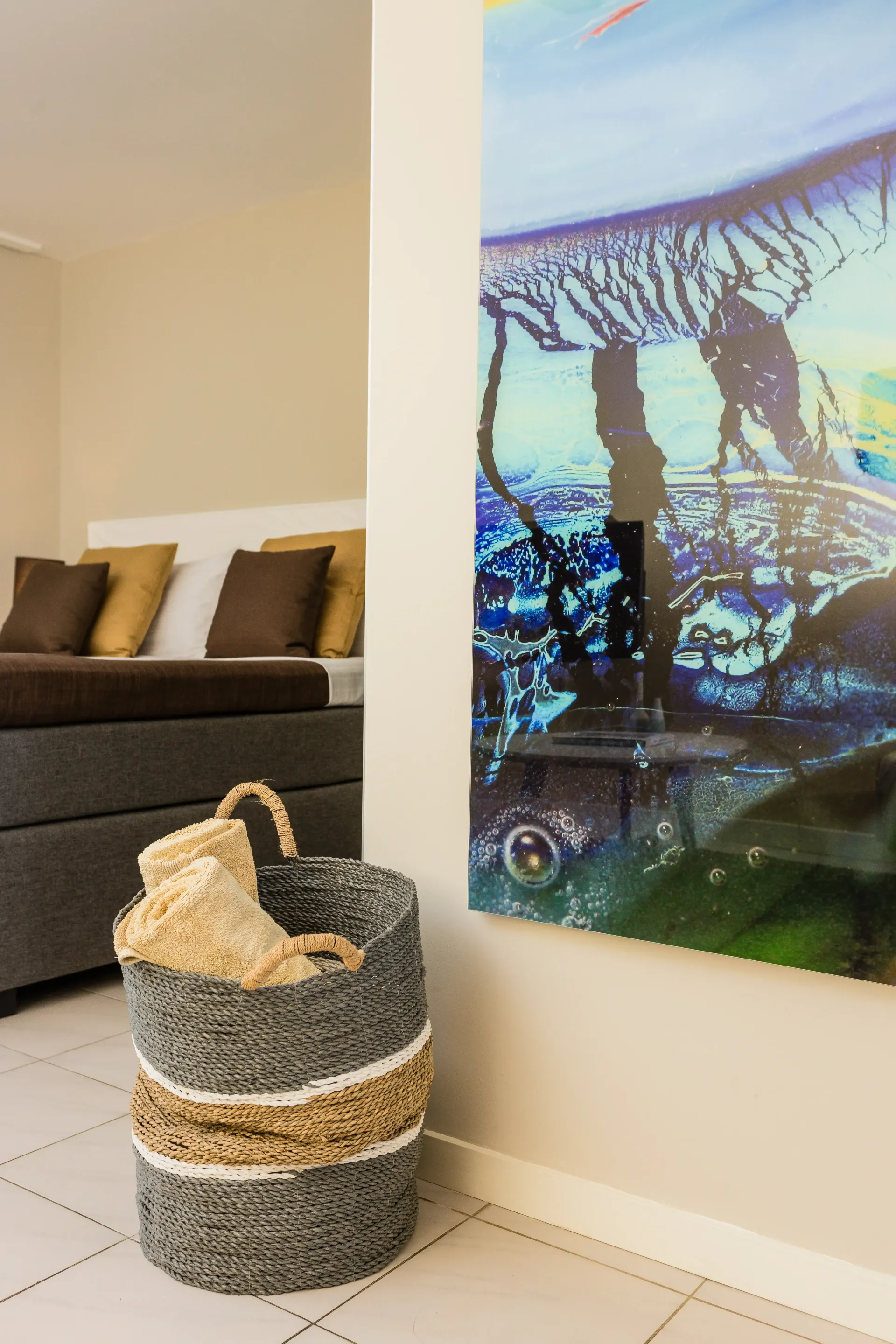 Woven basket on tiled floor, near a bed with brown and gold pillows and a colorful wall art.