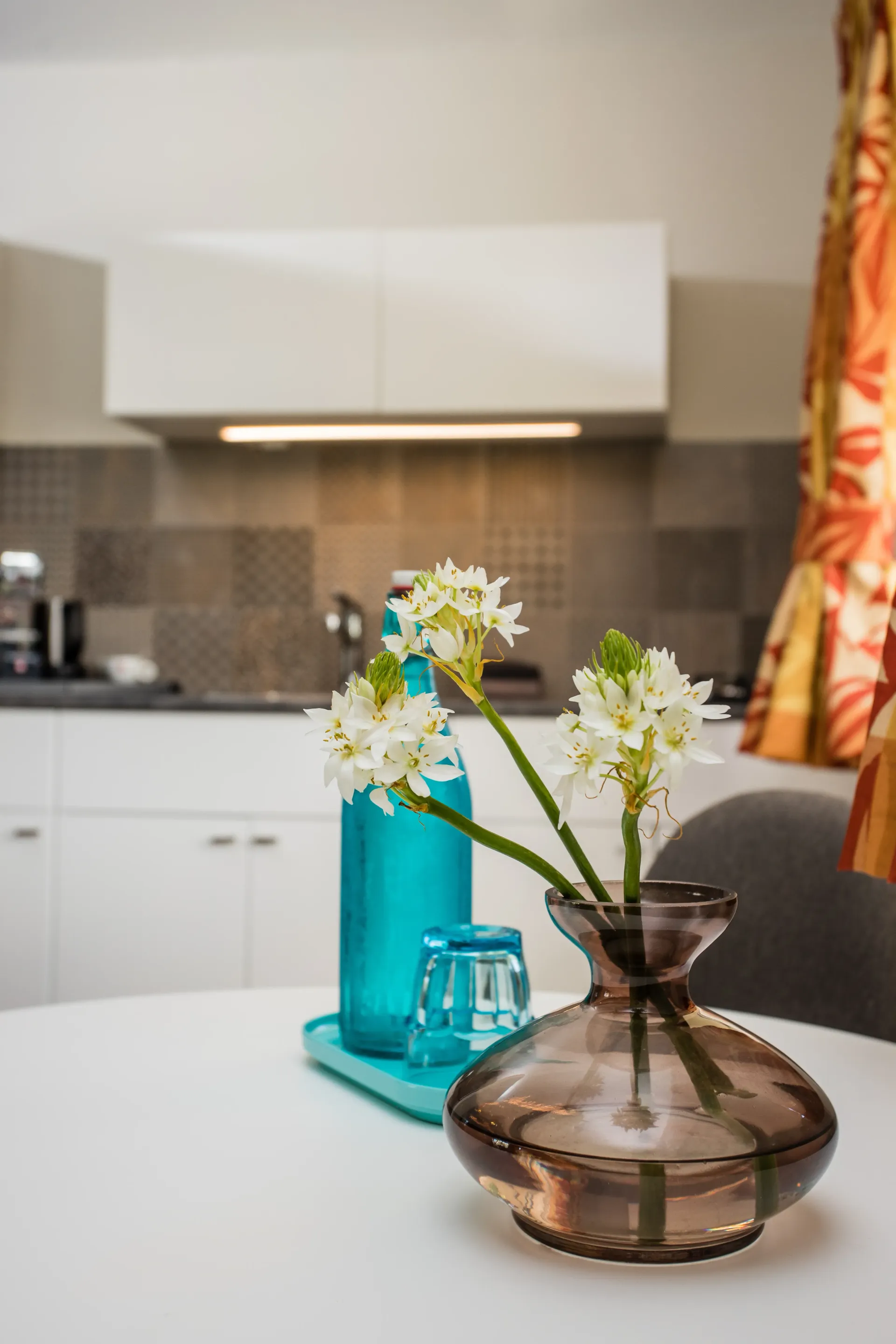 White flowers in a brown vase on a white table. A blue bottle and glass are behind it, in front of a kitchen.