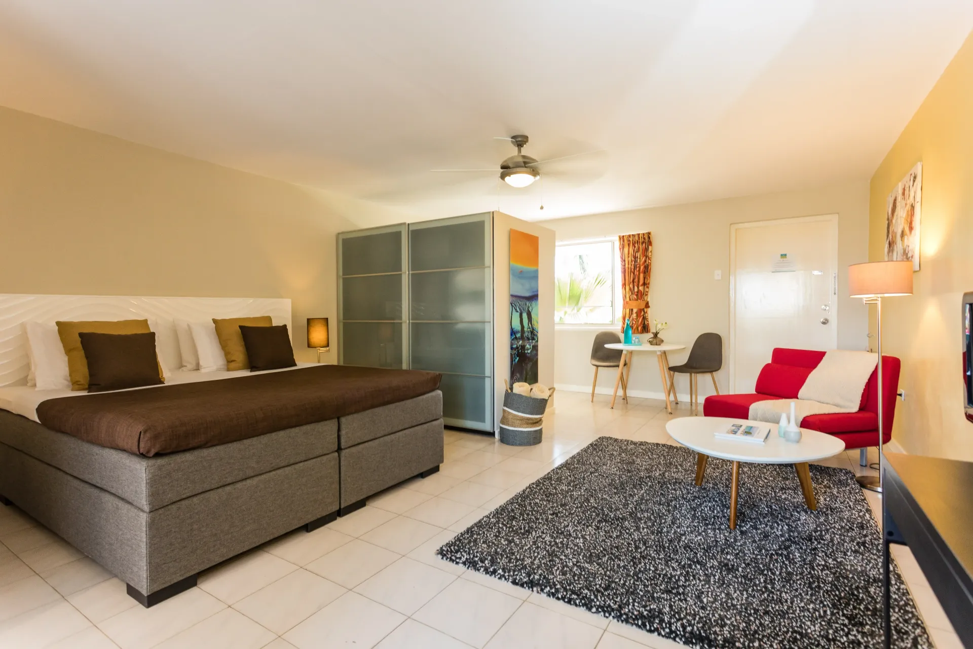 Hotel suite with king bed, seating area, and sliding glass closet. White walls and tile, neutral colors.