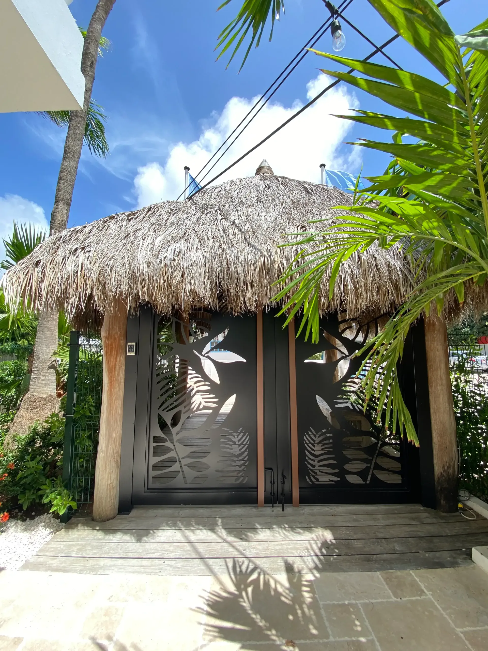 Tiki hut entrance with black metal gate featuring leaf cutouts, thatched roof, and concrete path.