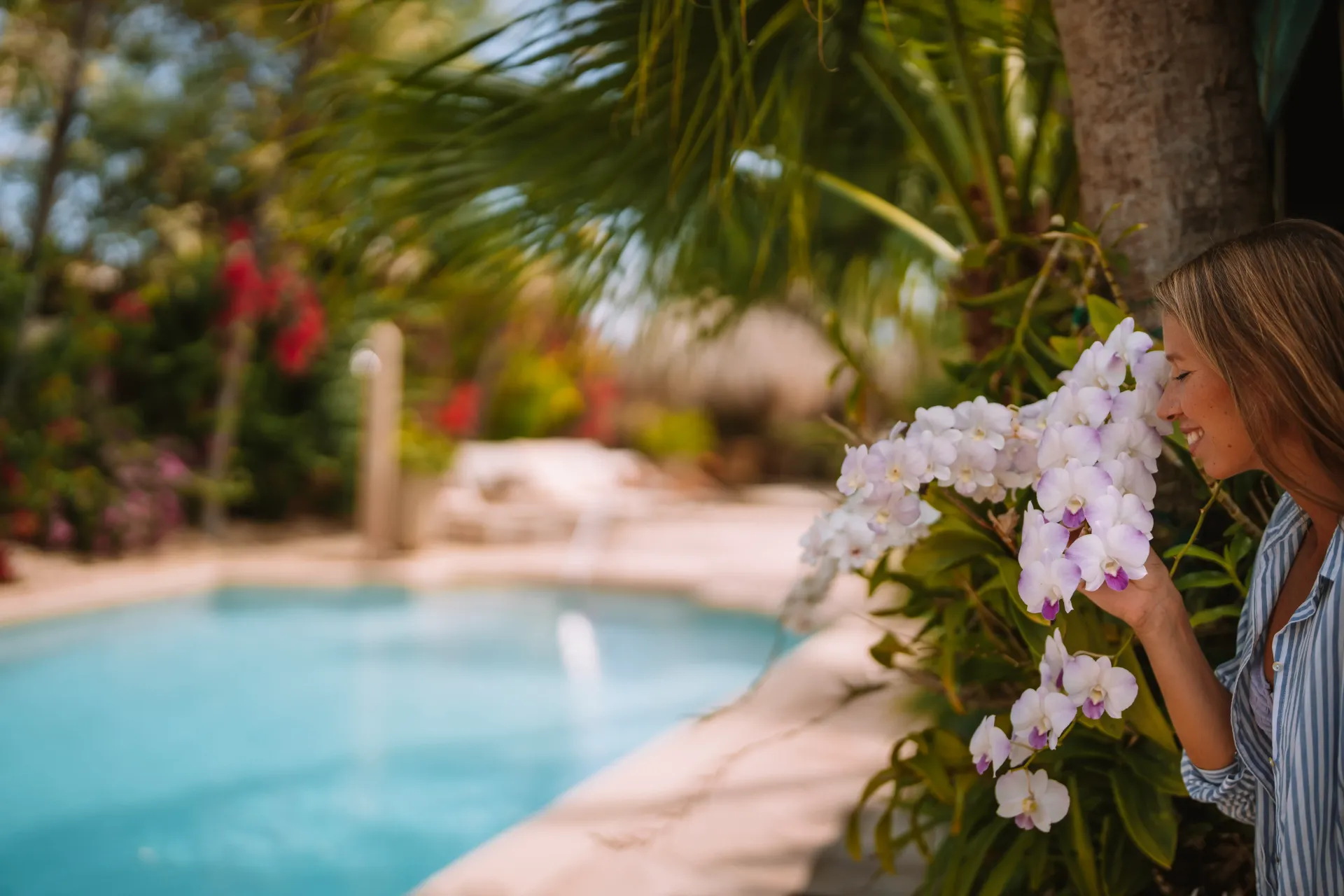 Woman smelling white orchids near a pool. Palm trees and red flowers in the background.