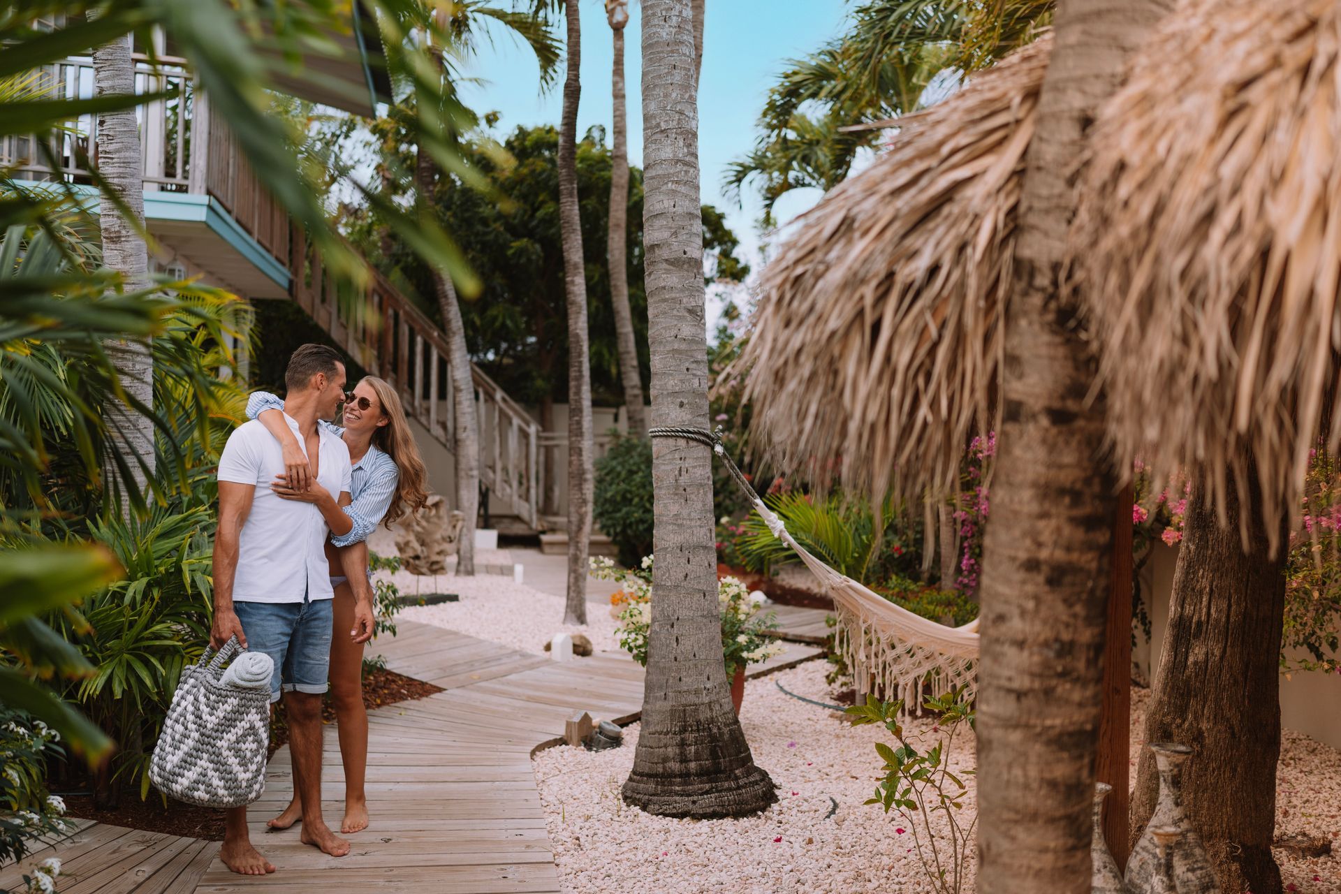 Couple hugging on a wooden path in a tropical resort, trees and a hammock are visible.
