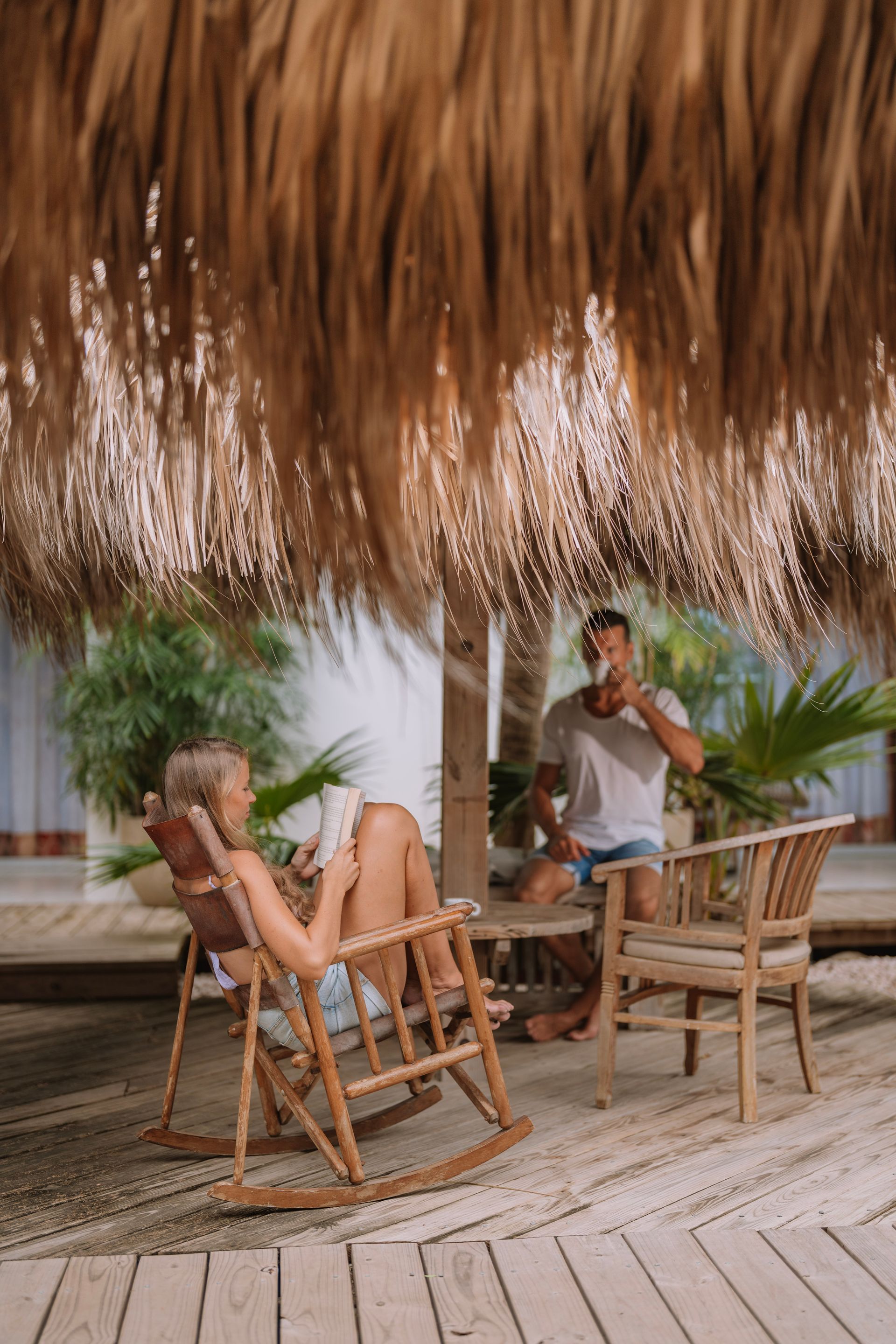 Couple relaxing outdoors under a thatched roof. Woman in a rocking chair, reading. Man drinks from a cup.