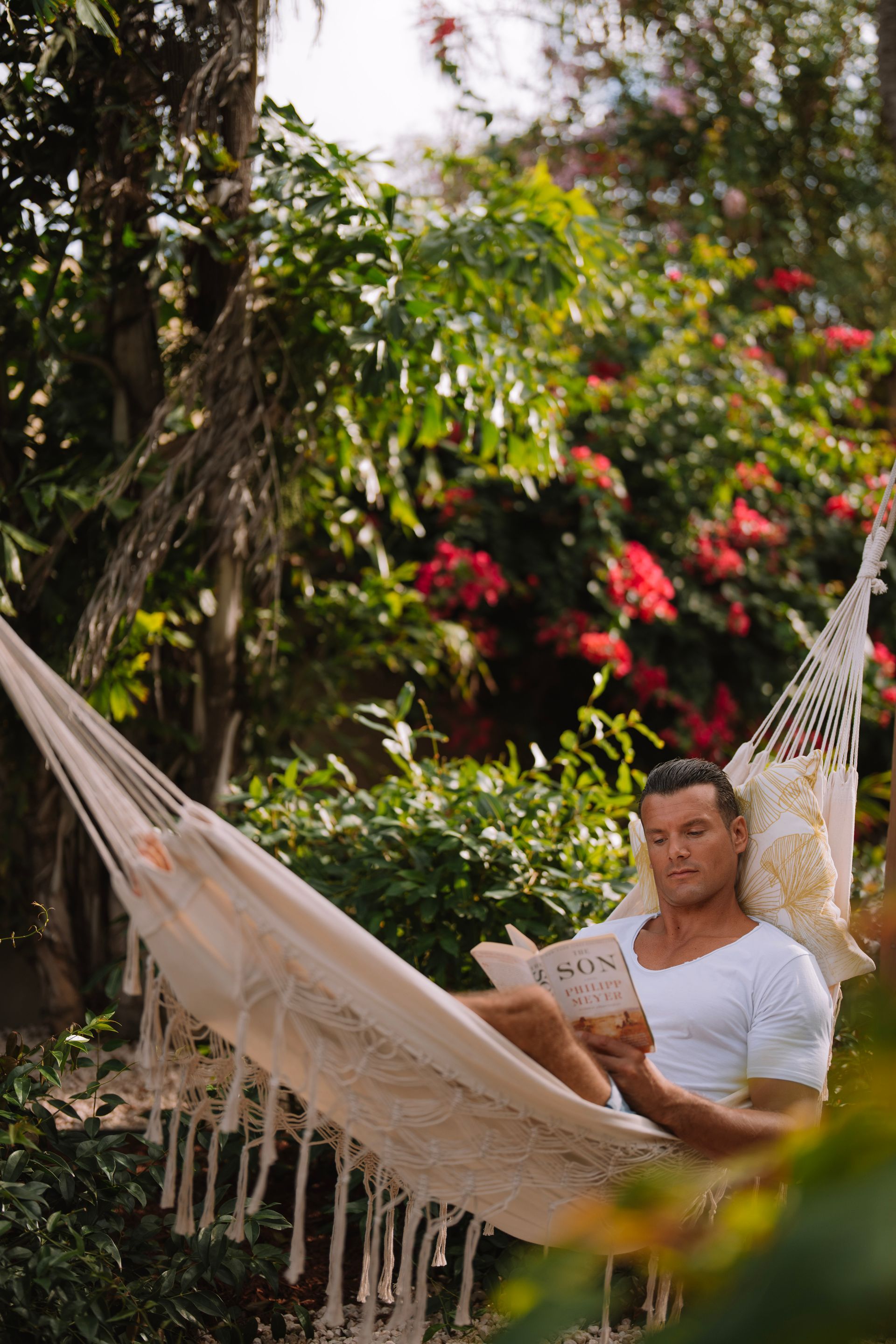 Man reading book in a hammock, surrounded by greenery and flowers.