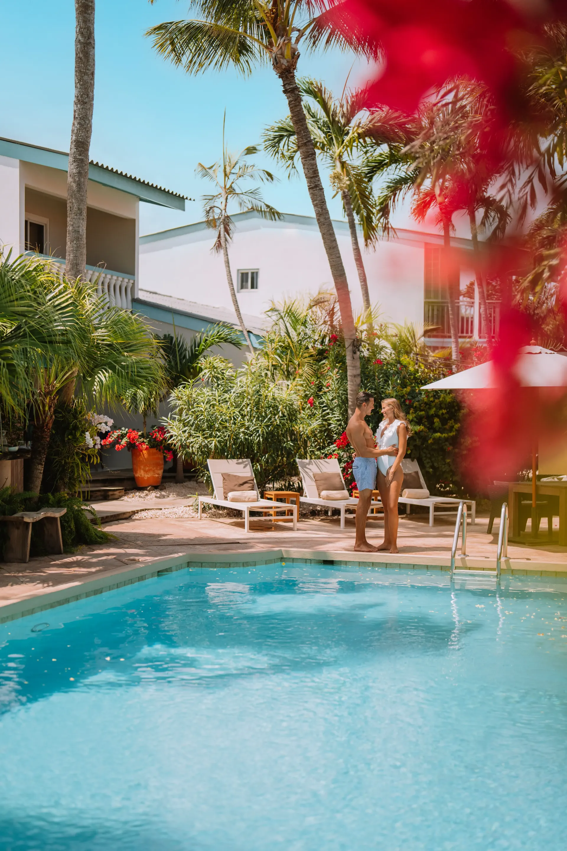 Couple standing by a pool in a tropical setting with white buildings, palm trees, and bougainvillea.