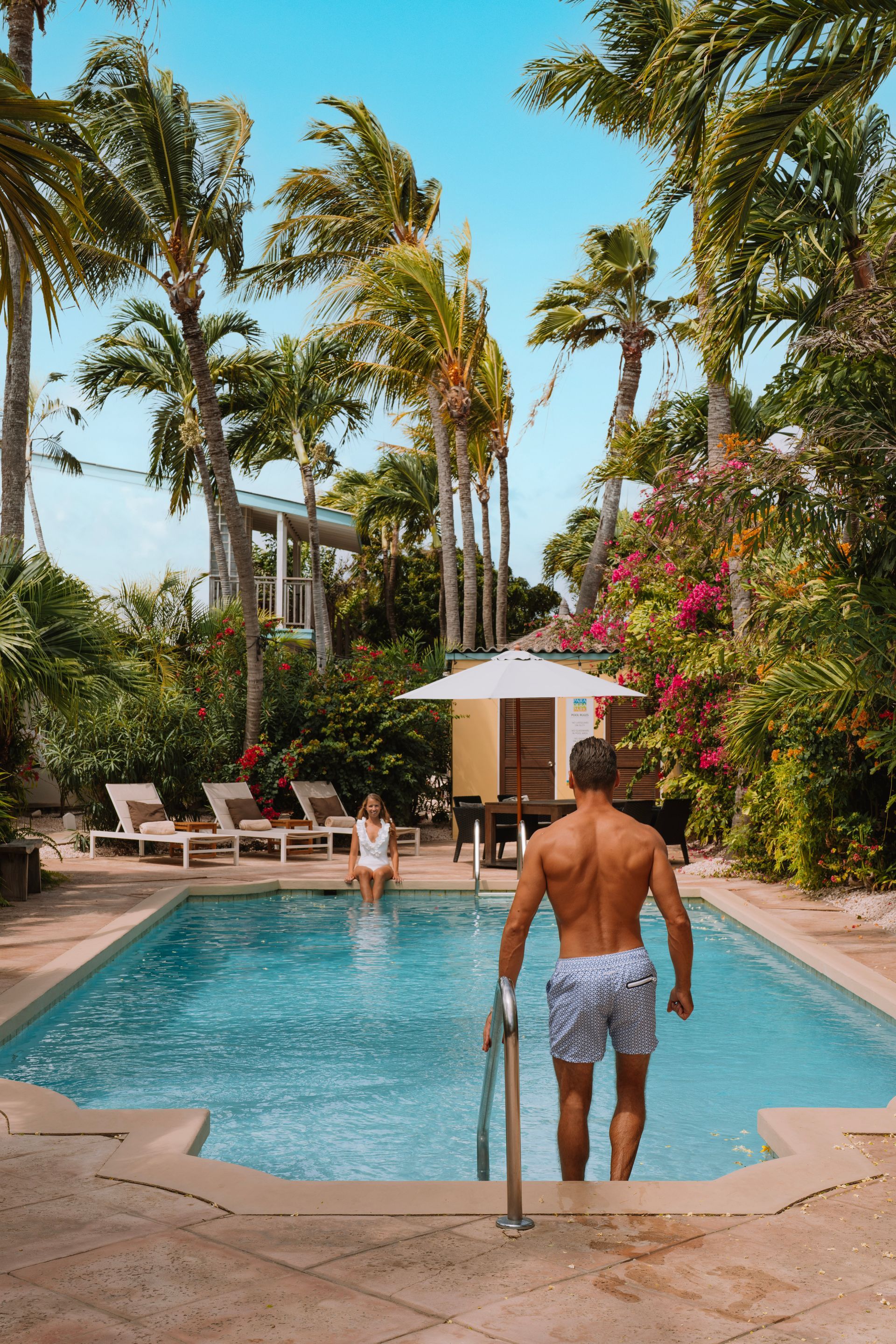 Man exiting pool, back view. Woman sitting poolside. Tropical setting with palm trees, blue sky.
