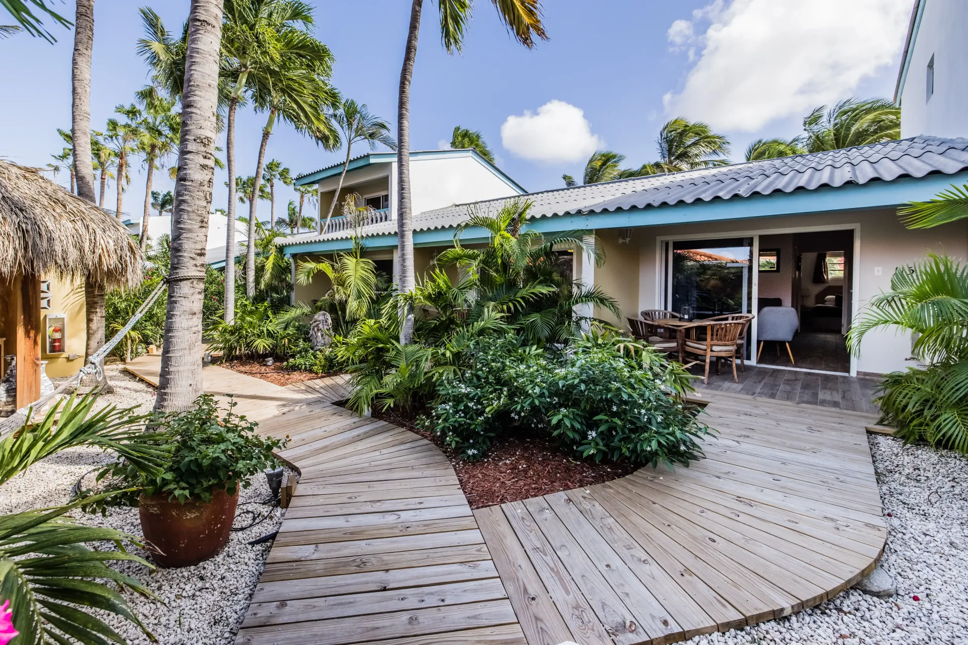 Wooden walkway leads to a building with a veranda, lush greenery, and palm trees in a tropical setting.