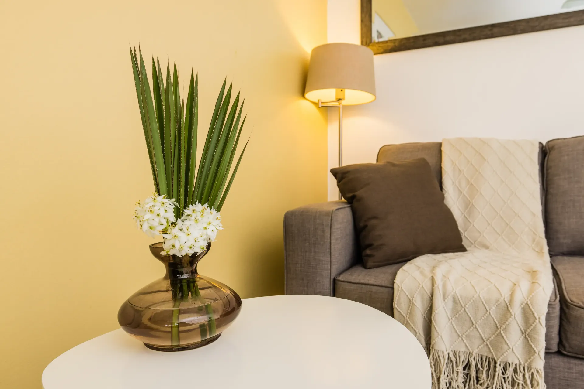 White table with vase of flowers, next to a gray couch with pillows, a blanket and a lamp.